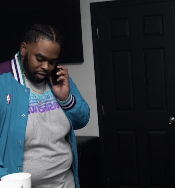 Man with braided hair talking on a cellphone, wearing a blue jacket and gray T-shirt, standing indoors.