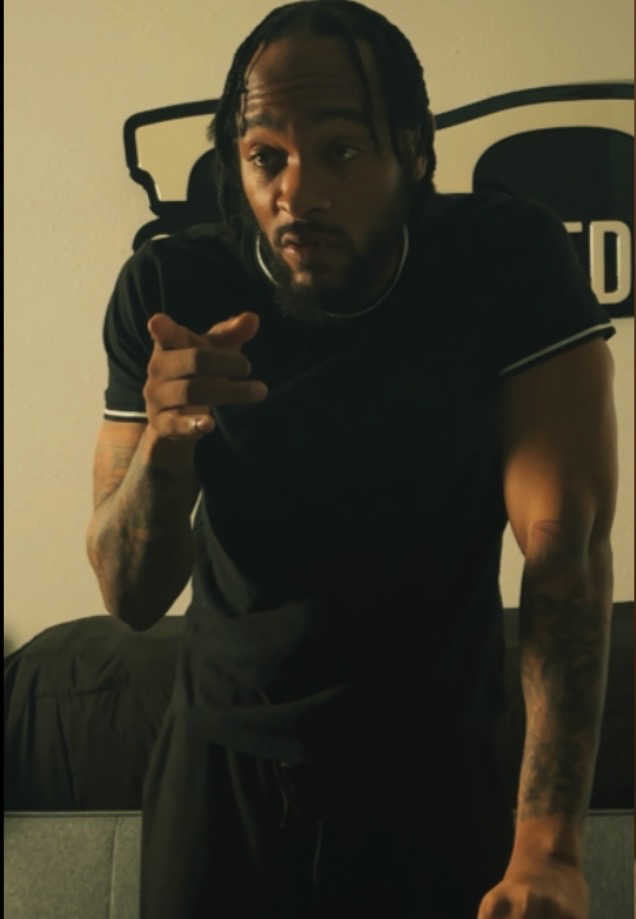 Man with braided hair pointing at the camera, standing indoors with a bed and wall art behind him.