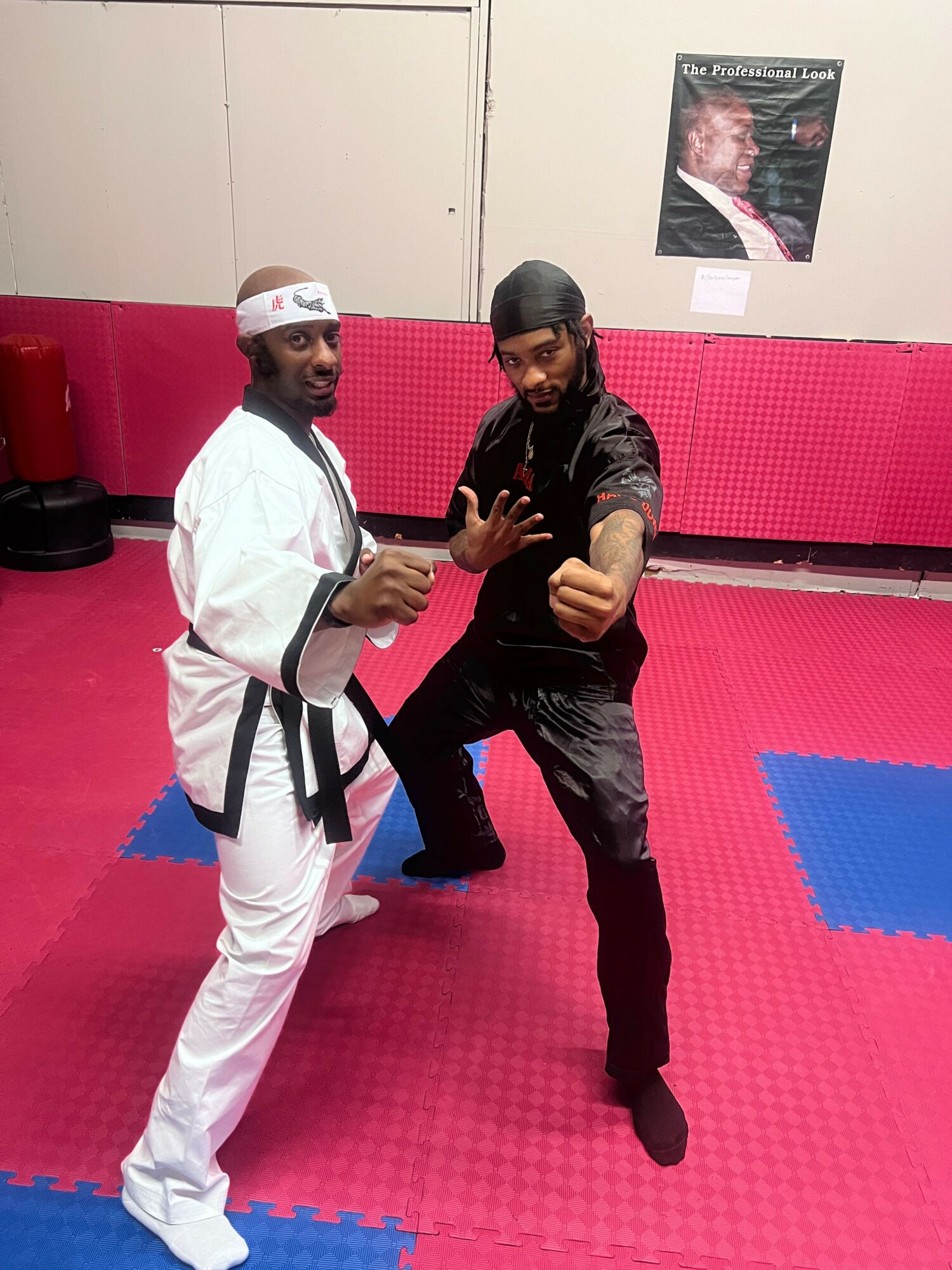 Two martial artists in uniform pose on a red and blue mat in a training room.
