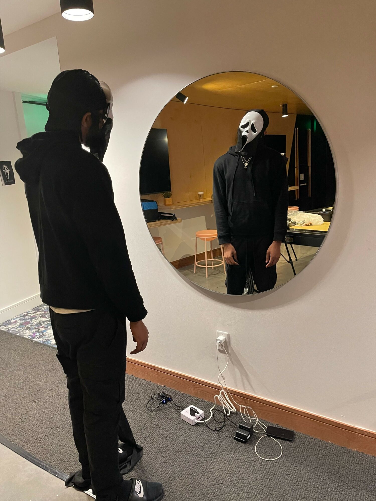 Person wearing a mask and black clothing looking at their reflection in a round mirror.