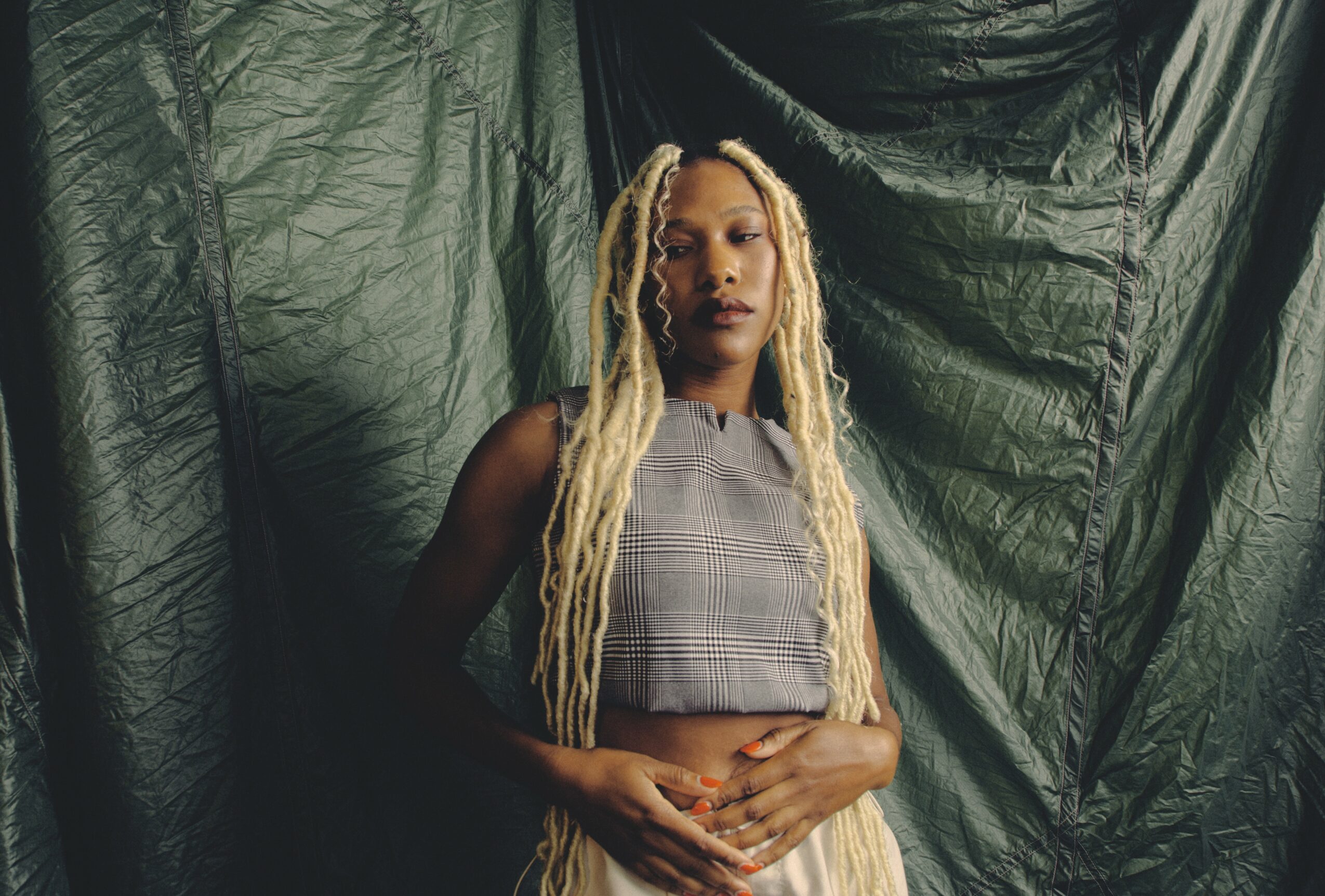 Woman with long blonde dreadlocks stands against dark fabric backdrop, wearing a sleeveless checkered top, hands clasped in front.