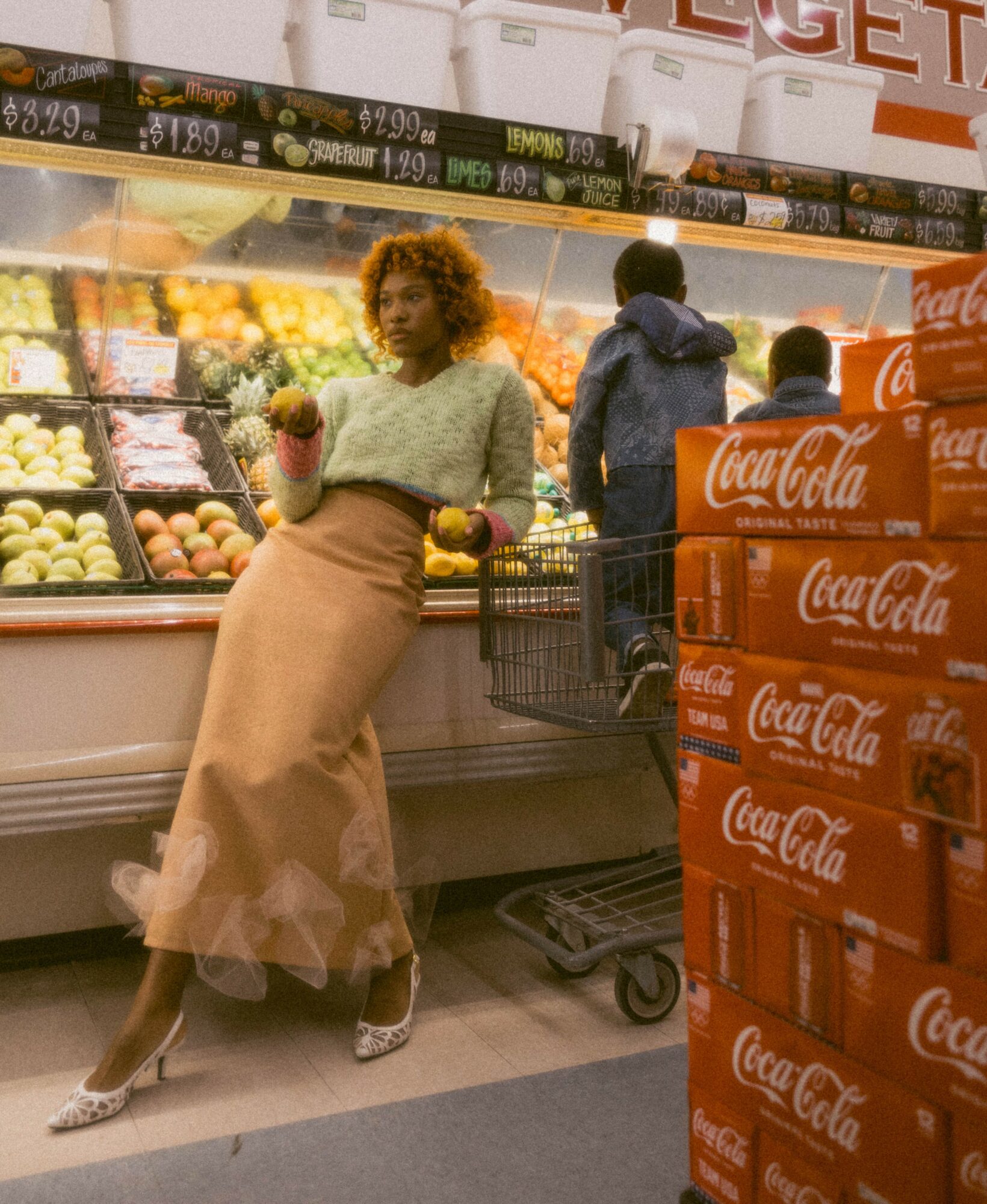 Woman in a grocery store holding a fruit, with a shopping cart and shelves of produce and Coca-Cola boxes nearby.