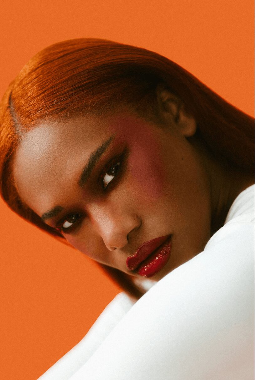 Close-up of a woman with red hair, makeup, and red lipstick against an orange background.