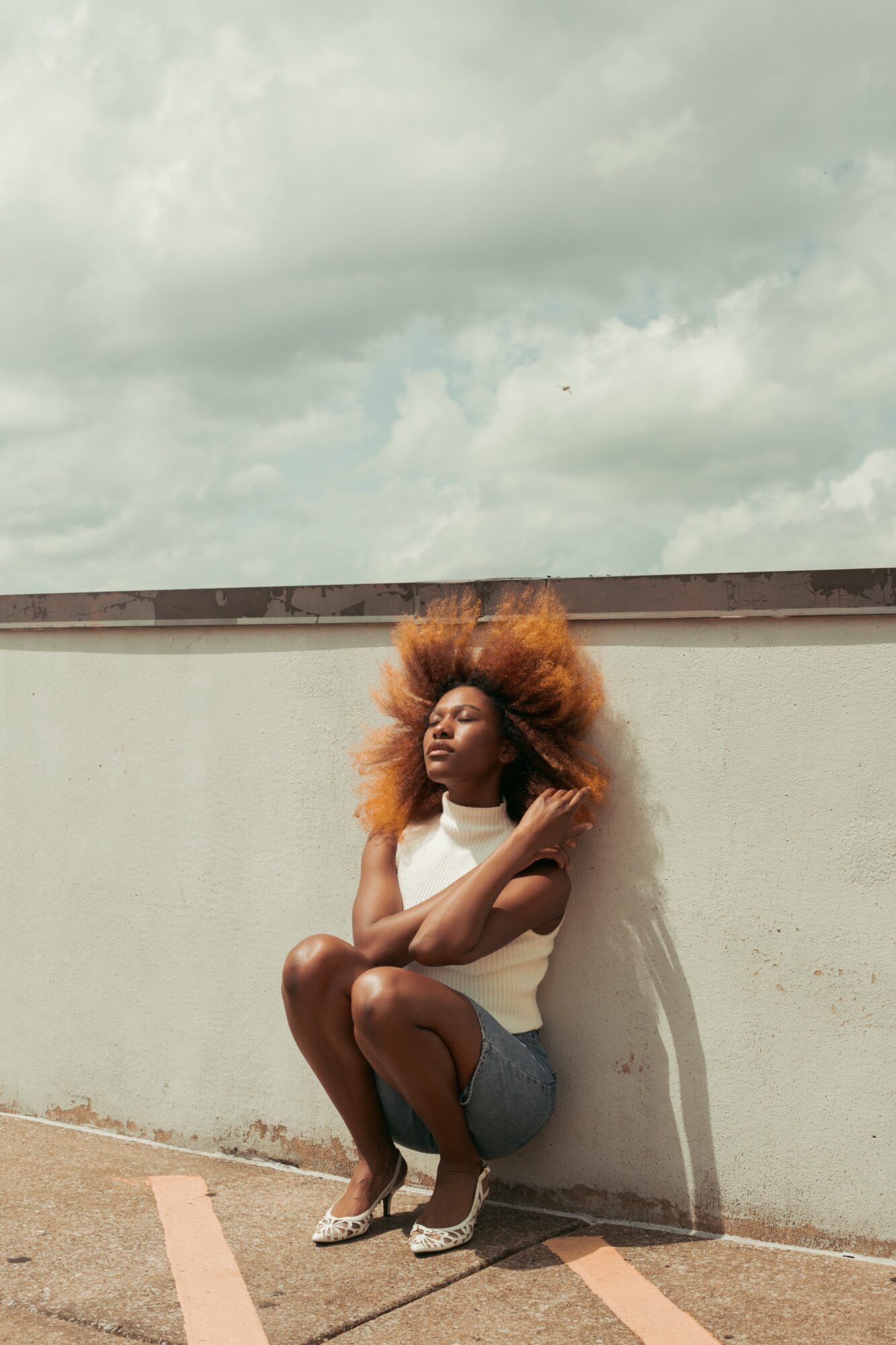 Person with large, curly hair sitting on the ground against a white wall outdoors, with cloudy sky above.