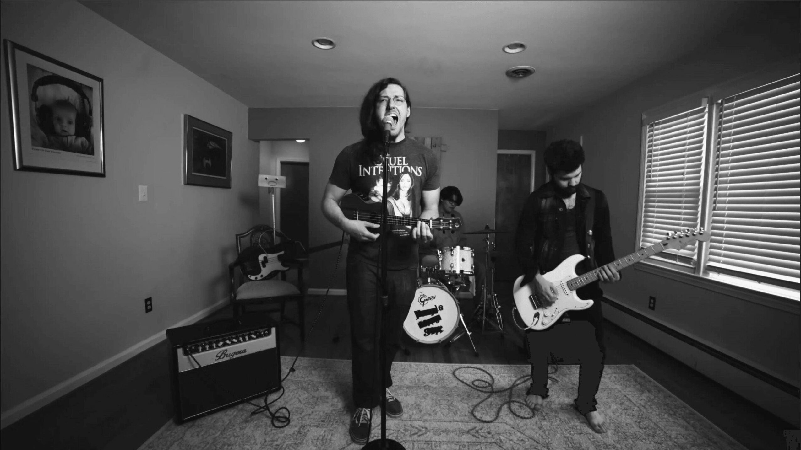 Band performing in a room with three members: singer, guitarist, and drummer, with musical equipment and windows with blinds.