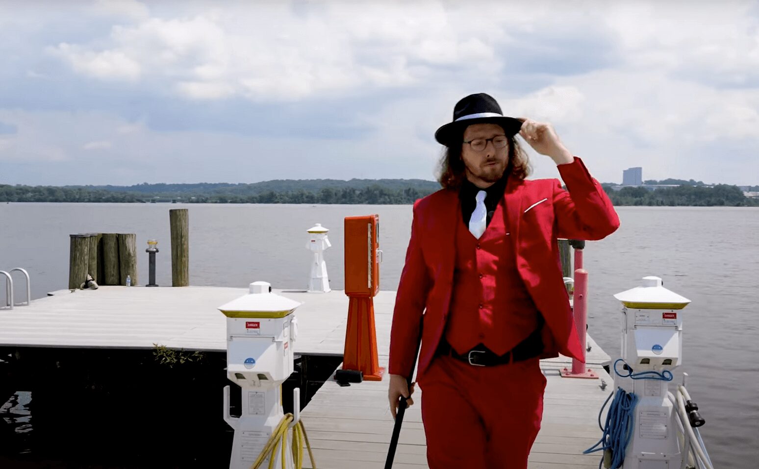 Person in red suit and black hat standing on dock near water, holding a cane and adjusting hat.