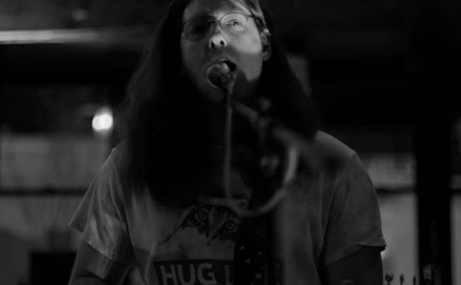 Person with long hair and glasses speaking into a microphone indoors.