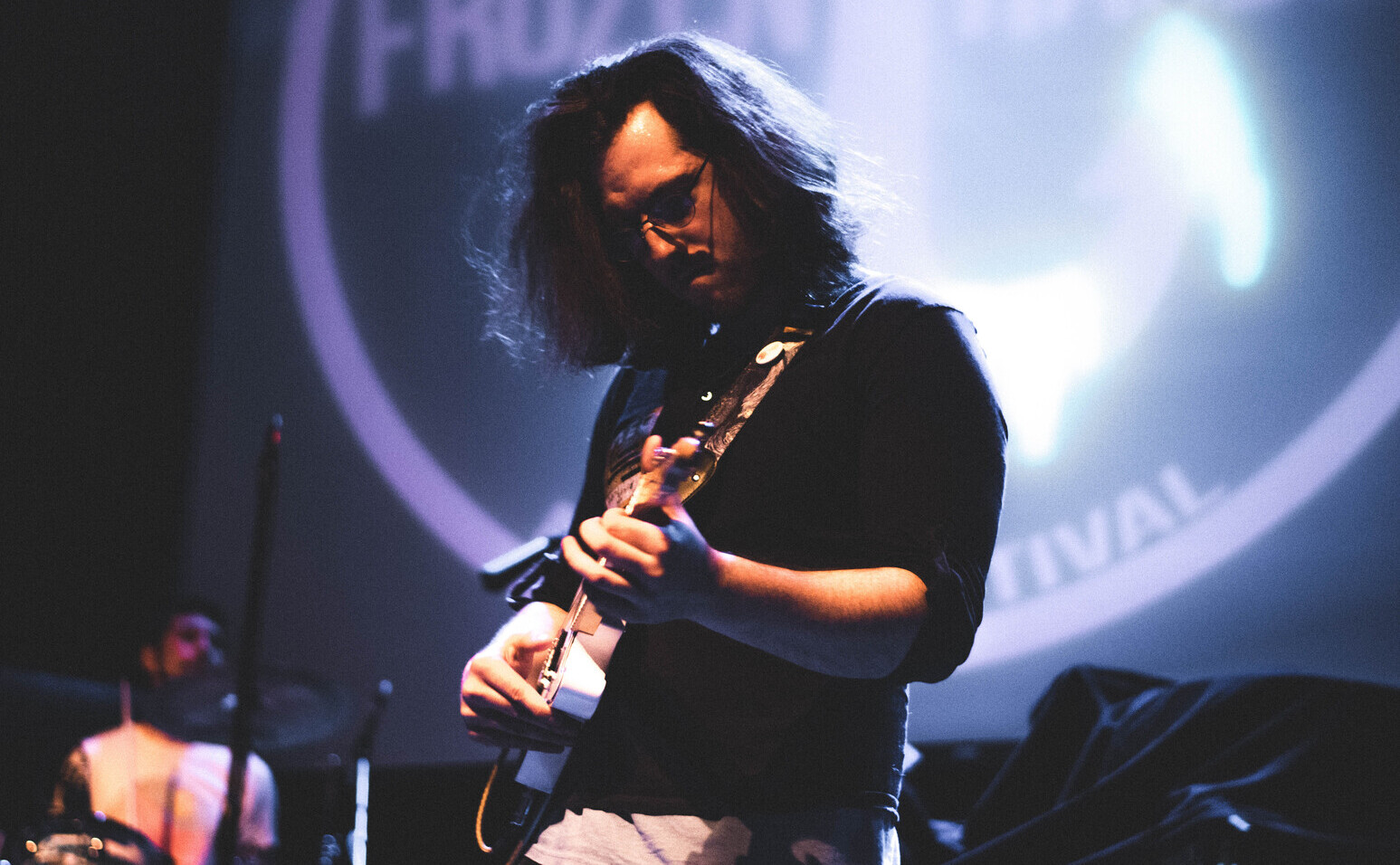 Person playing guitar on stage with a large circular logo in the background, stage lighting illuminating the scene.