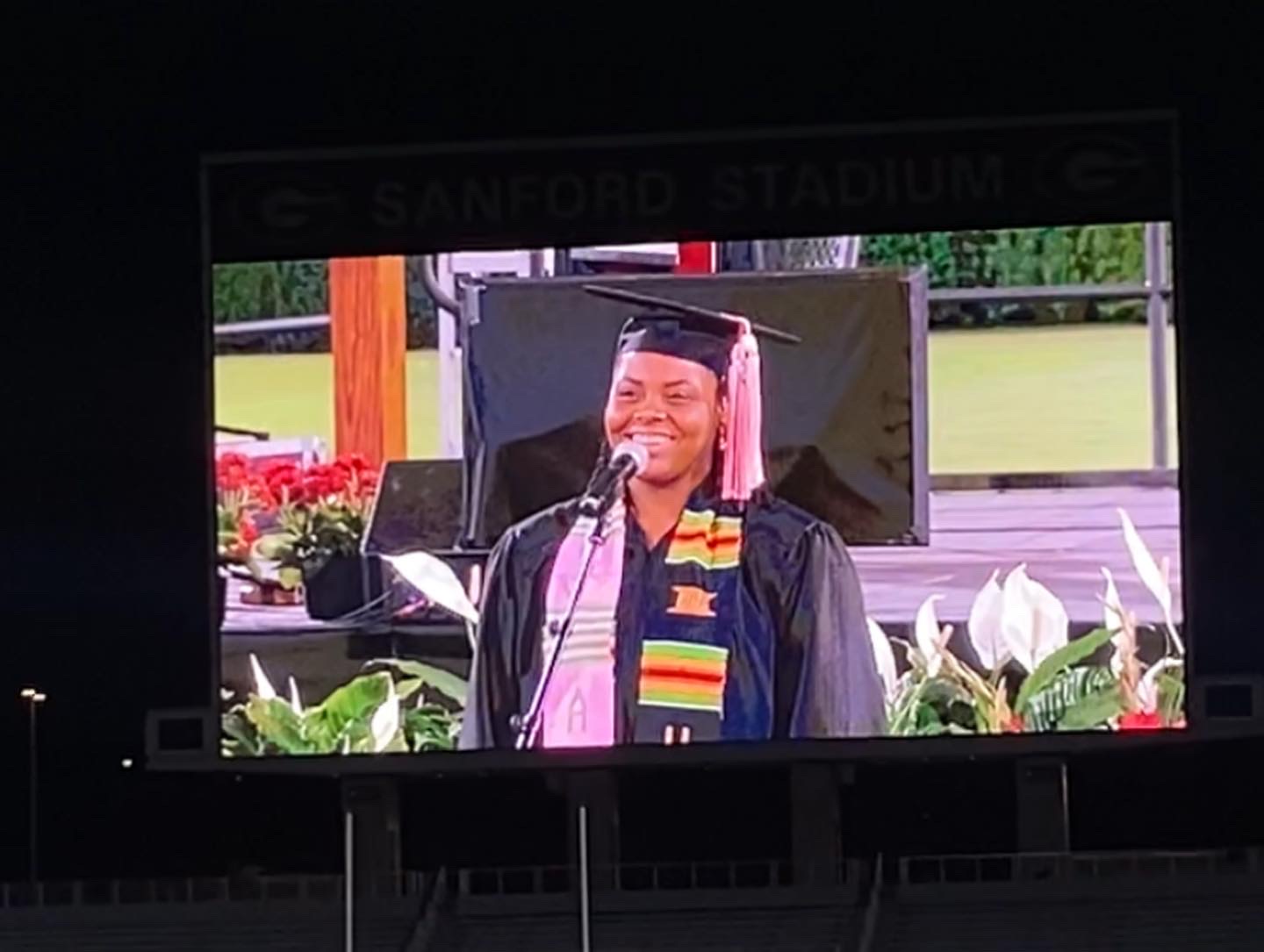 Person in graduation cap and gown smiling, standing outdoors with flowers and a screen behind them.