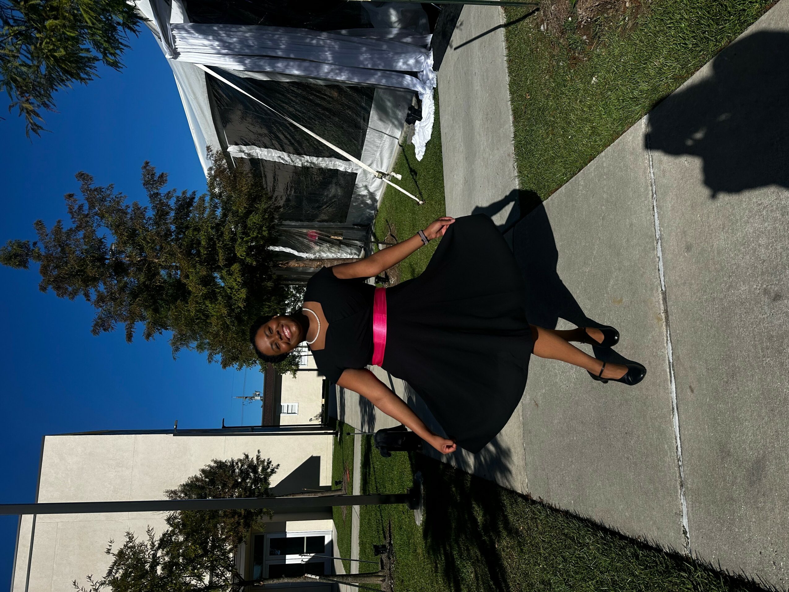 Woman in black dress with pink belt walking on sidewalk, holding a phone, with trees and tents in background.