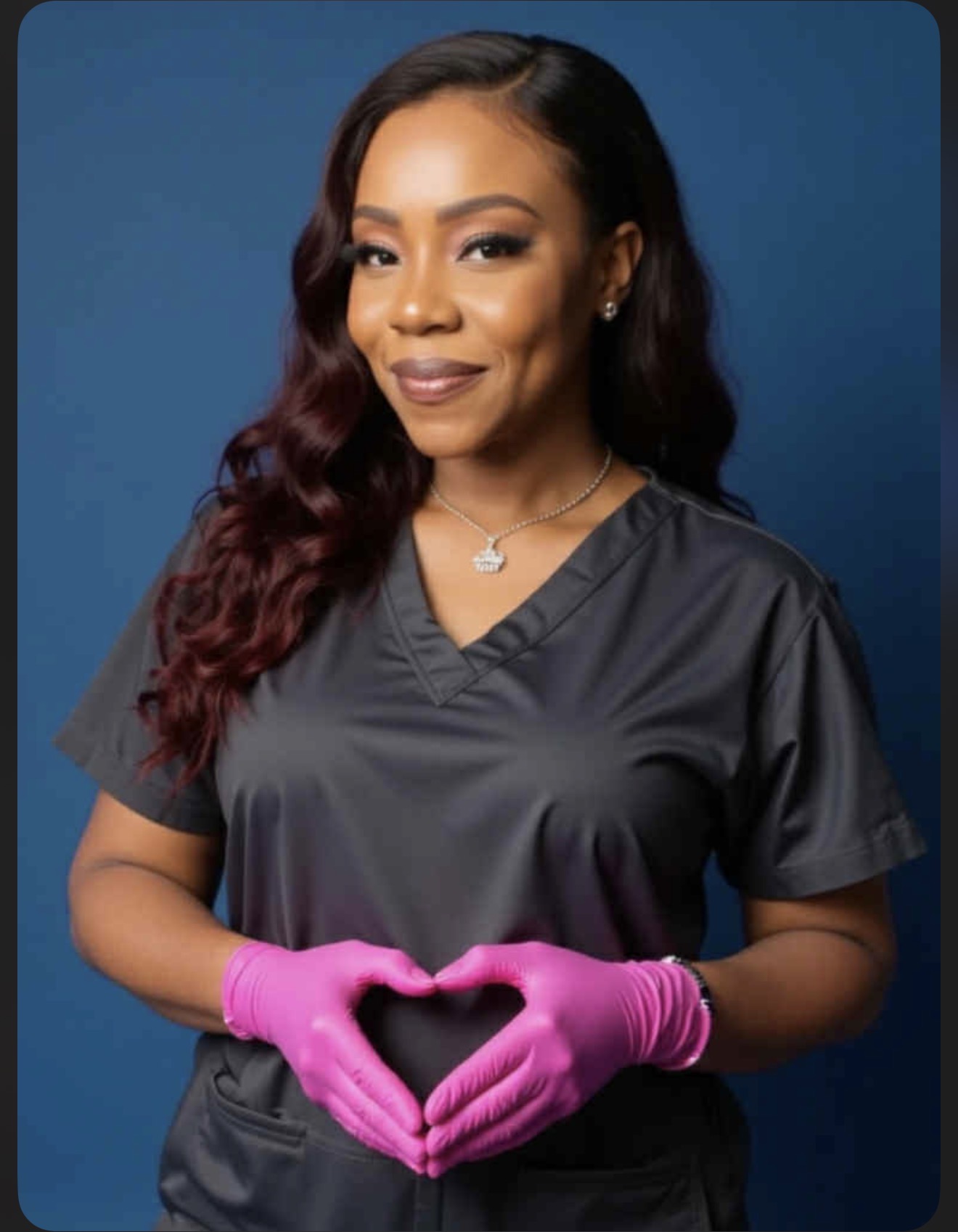 Woman in dark scrubs wearing pink gloves, making a heart shape with her hands, smiling, against a blue background.