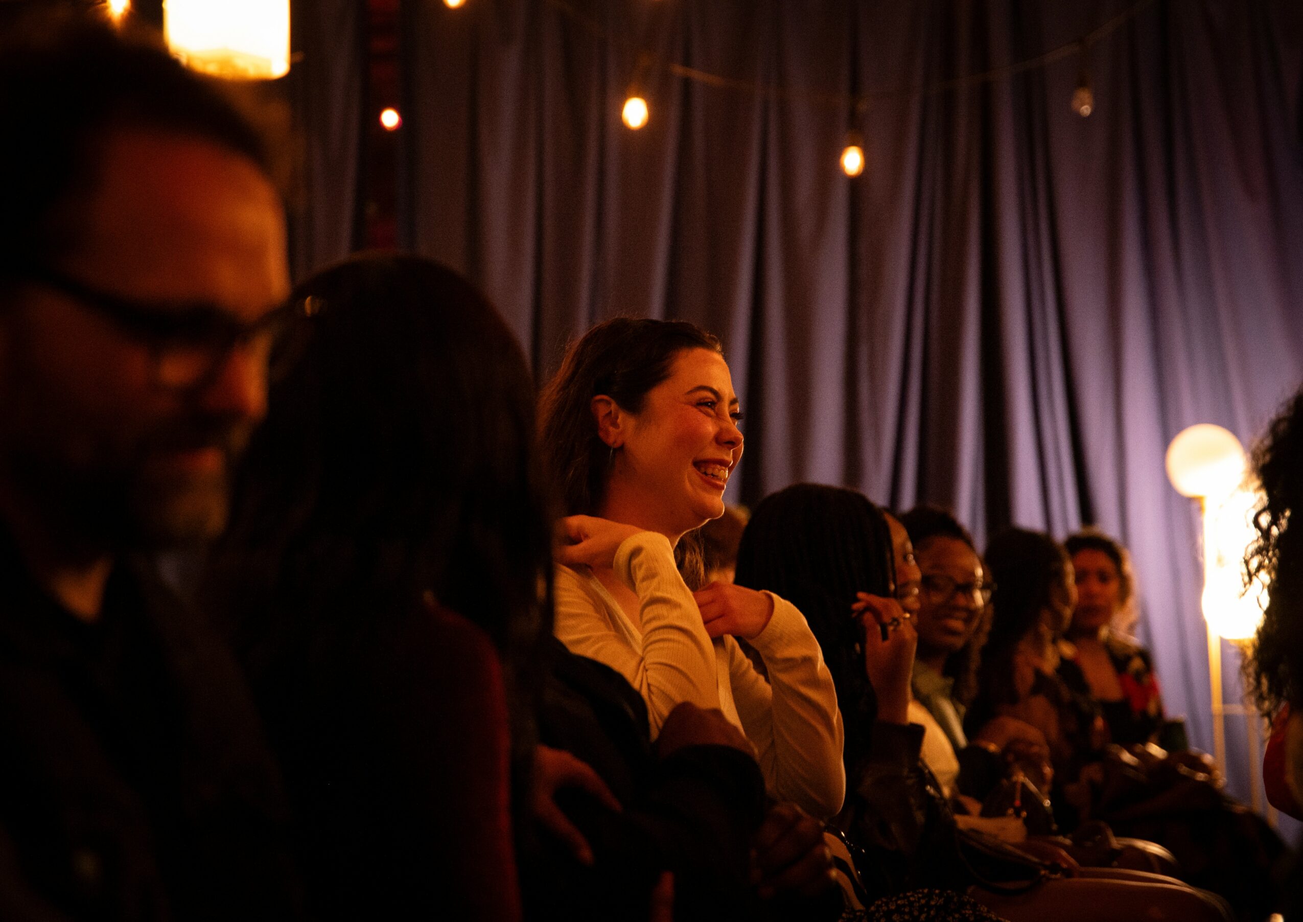 Group of people sitting indoors, smiling and engaging, with curtains and string lights in the background.