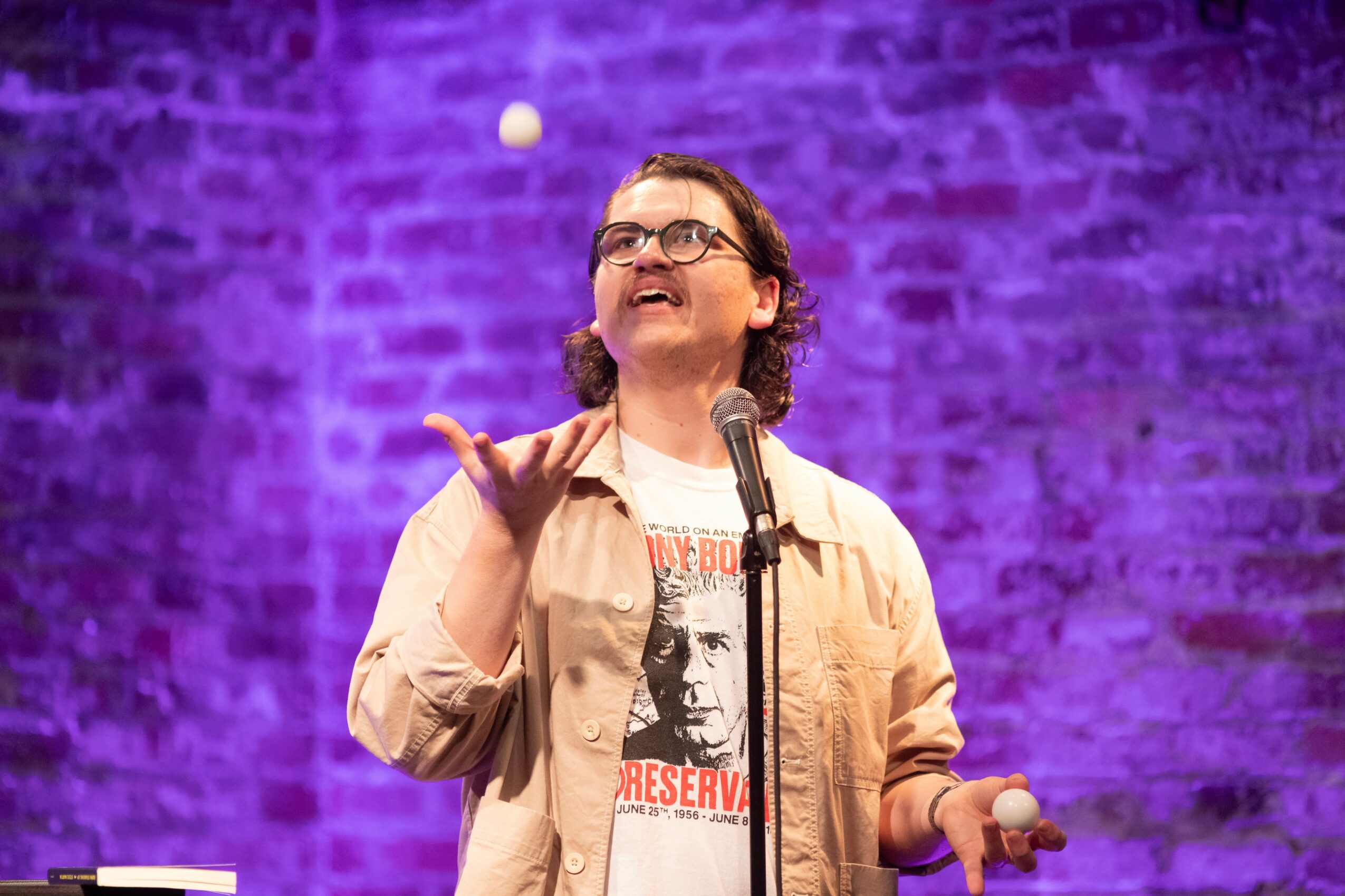 Person with glasses juggling a ball on stage with purple brick wall background.
