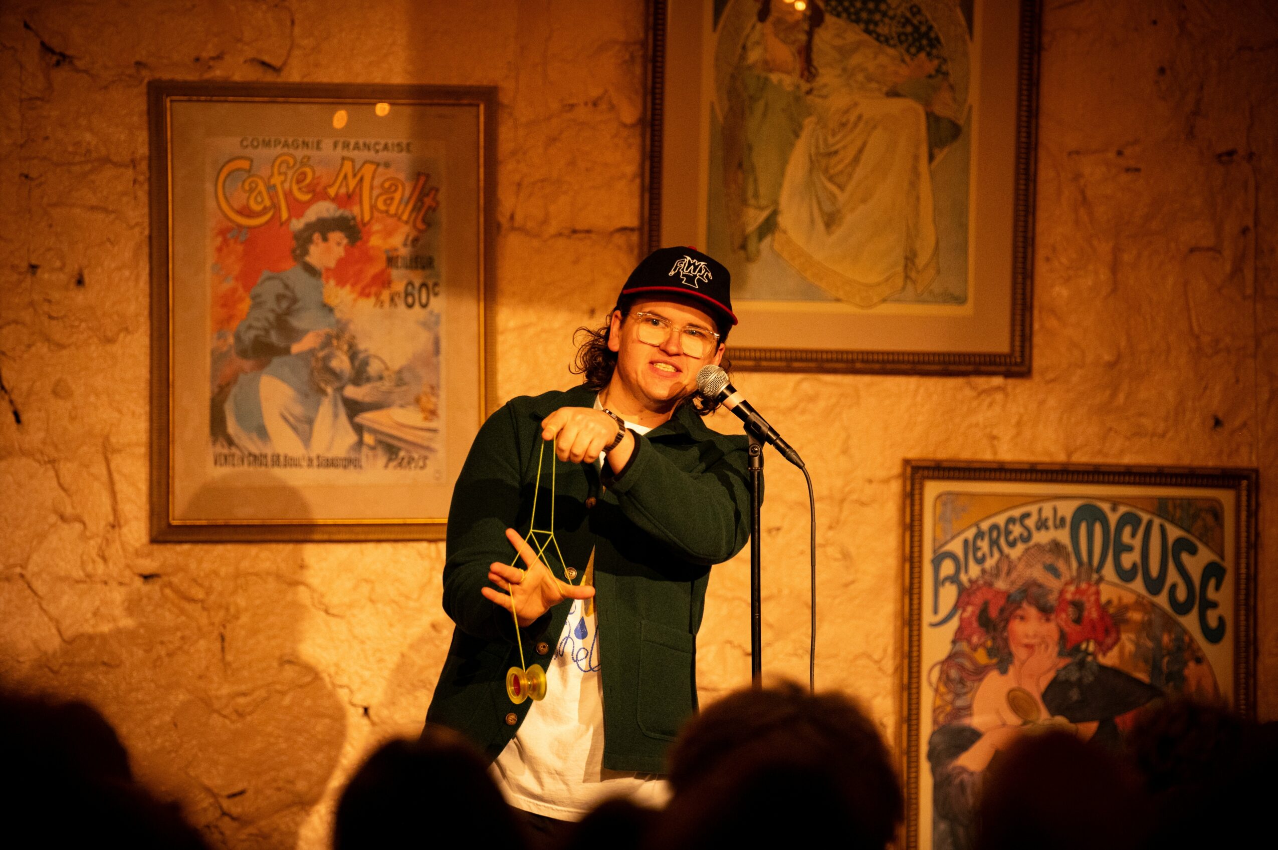 Person performing stand-up comedy on stage with framed posters on brick wall behind them.