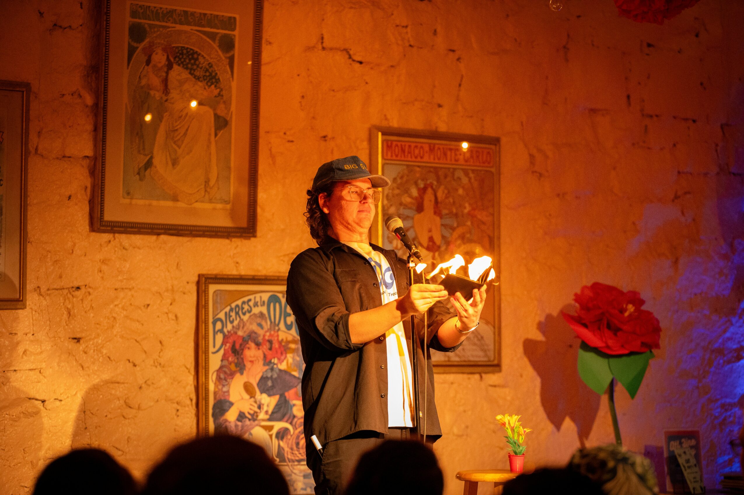 Person reading from a book on stage with framed artwork and a large red flower decoration behind them.