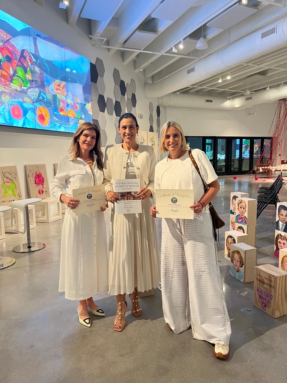 Three women in white dresses holding certificates, standing in an art gallery with colorful artwork and displays.