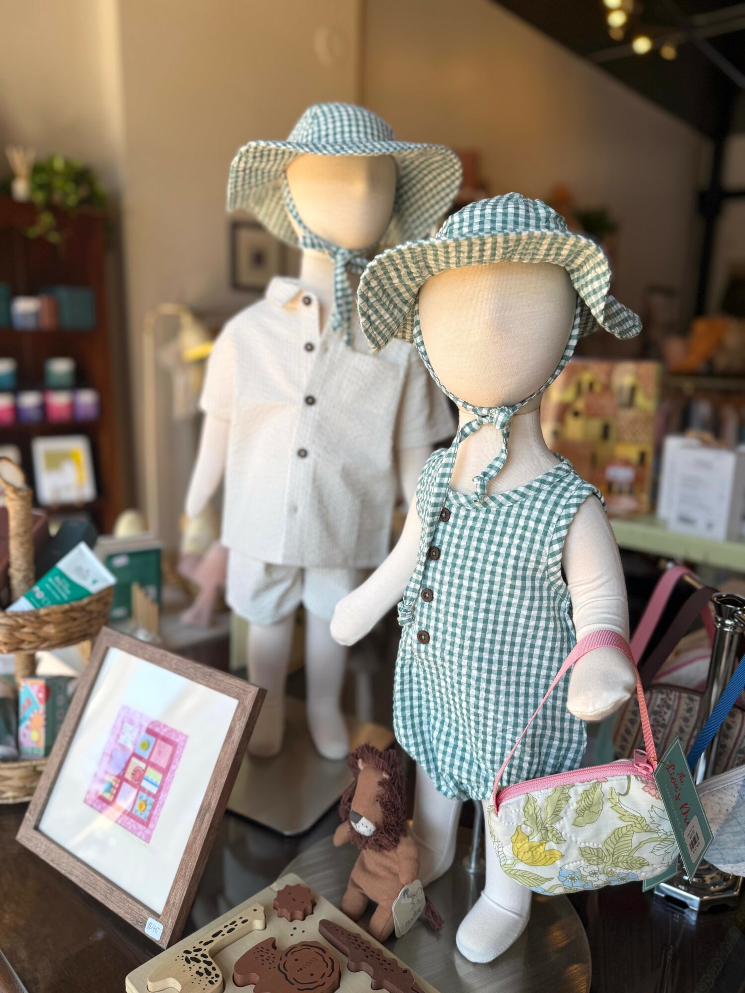 Two child mannequins dressed in summer outfits with hats, displayed in a store with framed pictures and accessories.