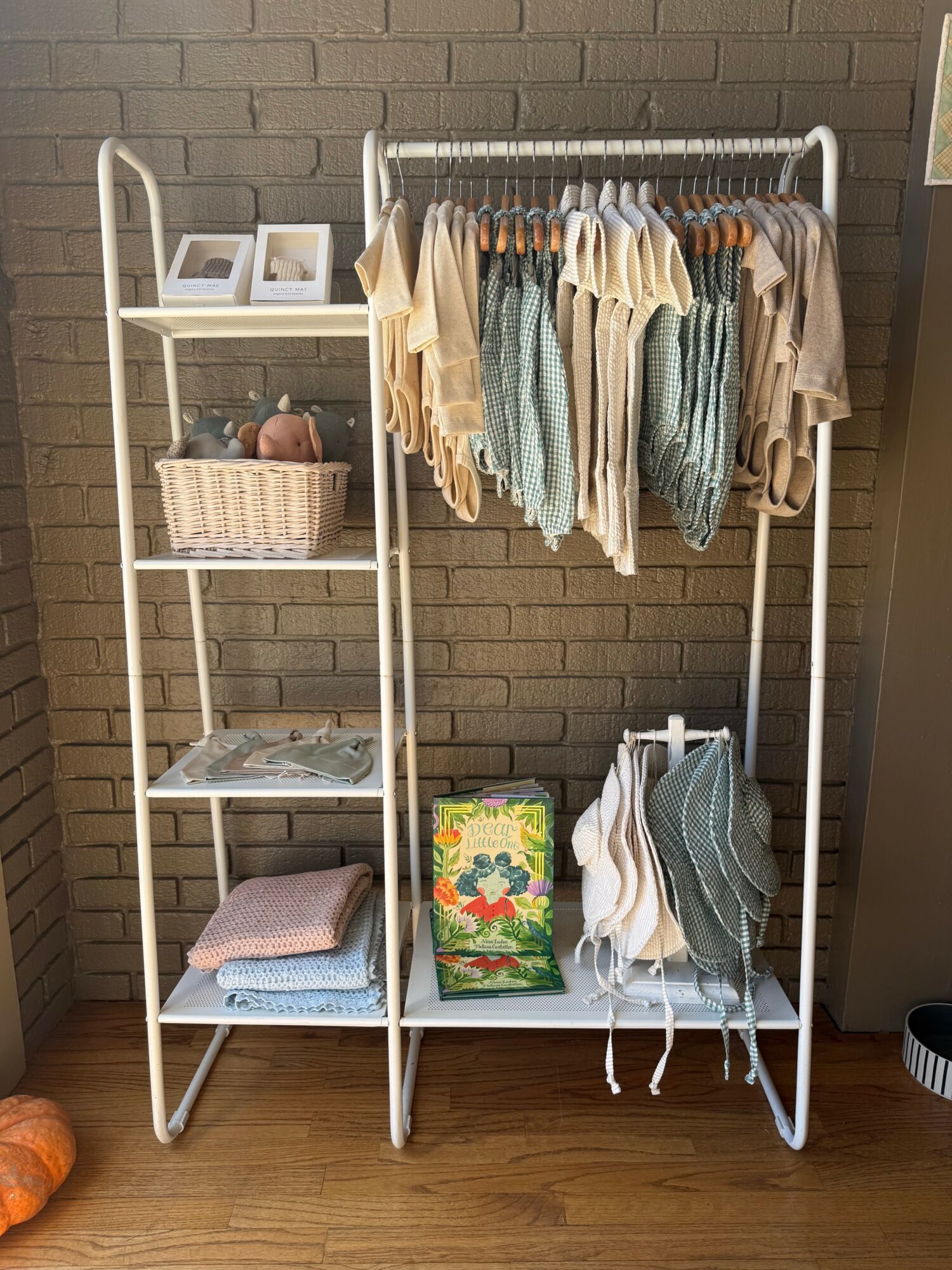 White clothing rack with hanging clothes, shelves with baskets, books, and folded towels, against a brick wall.