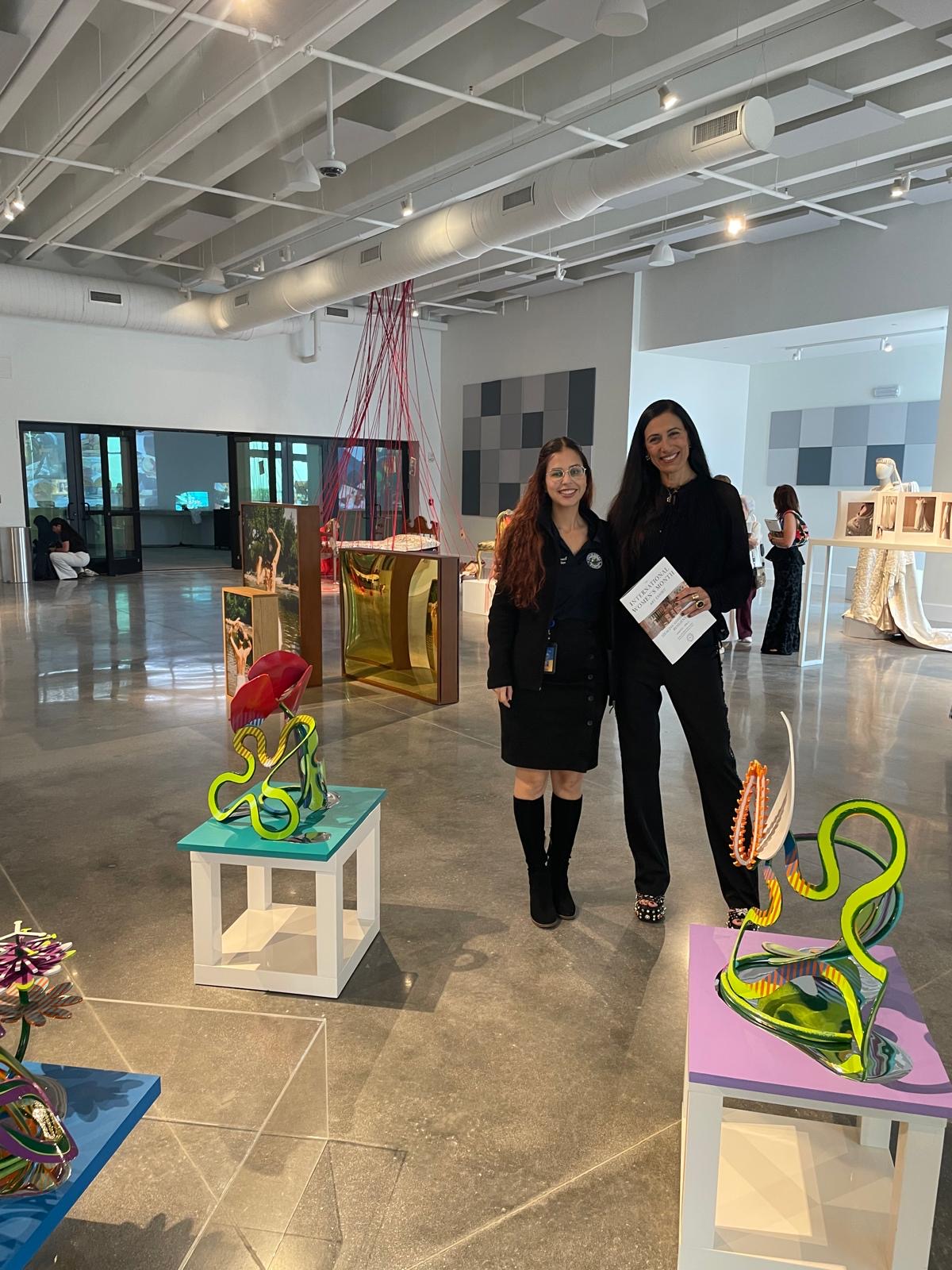 Two women stand in an art gallery, surrounded by colorful sculptures on display, with other visitors in the background.