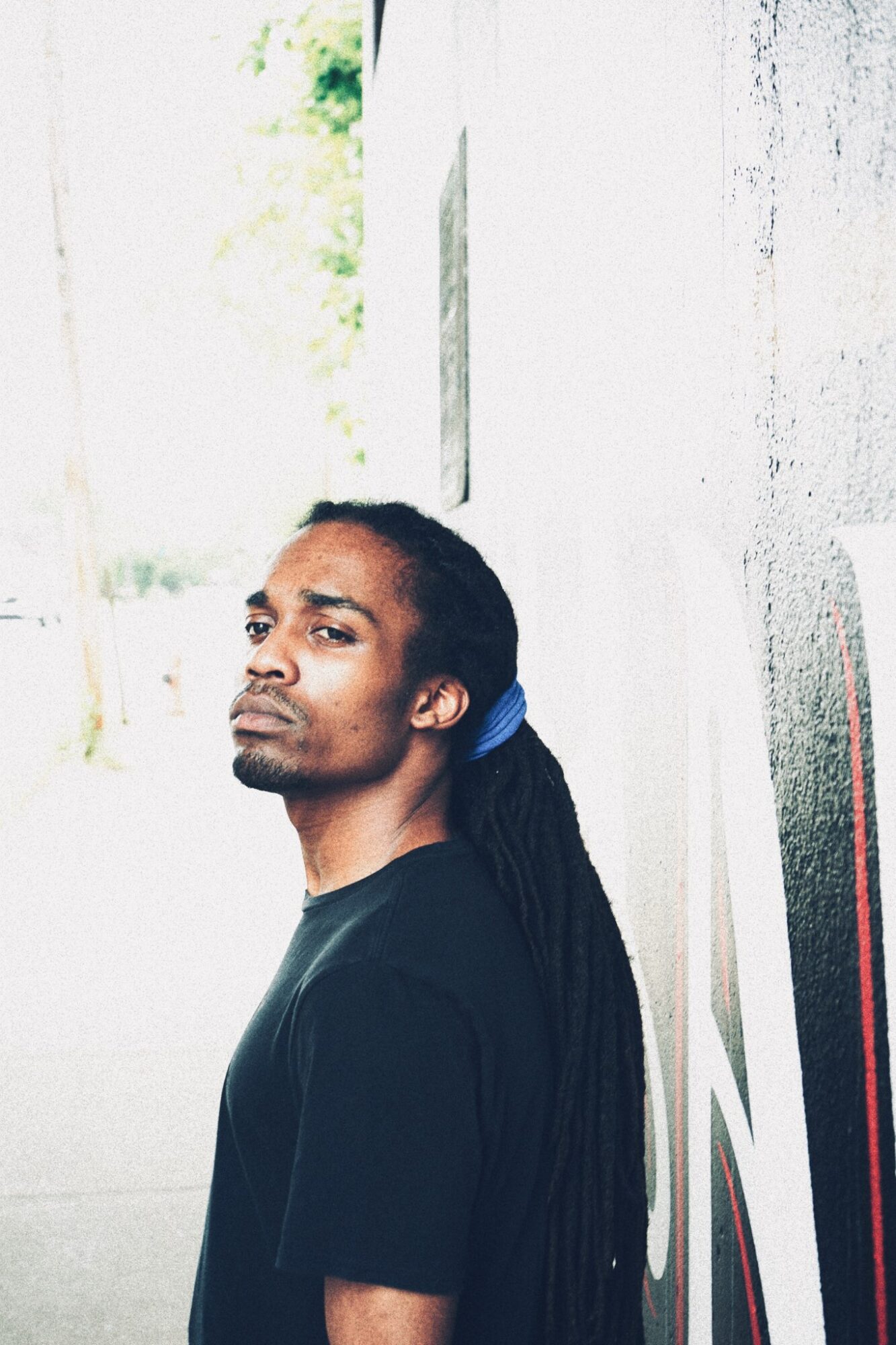 Man with long dreadlocks leaning against a white wall outdoors, looking at the camera.