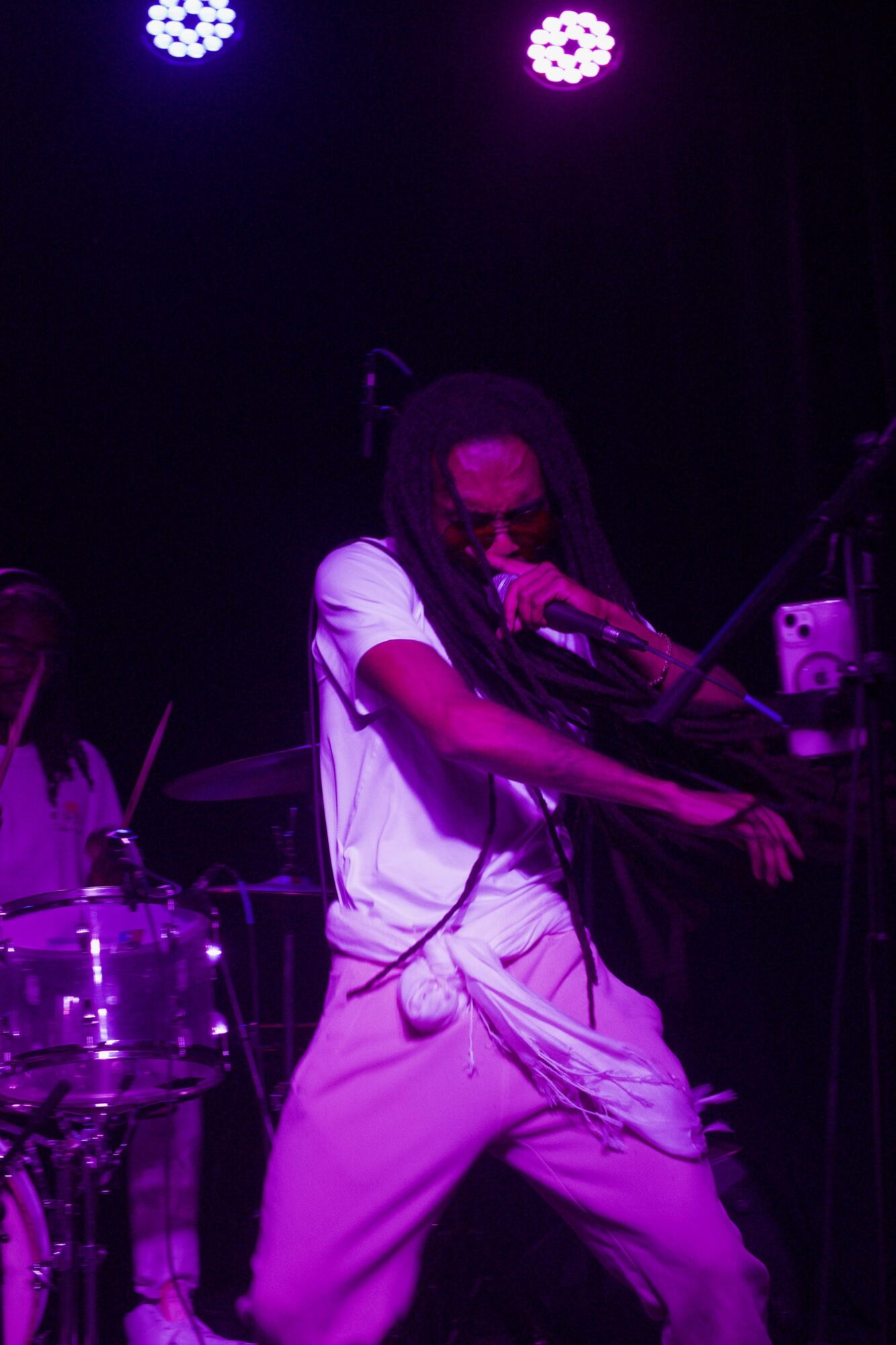 Performer with long hair singing into microphone on stage with purple lighting.