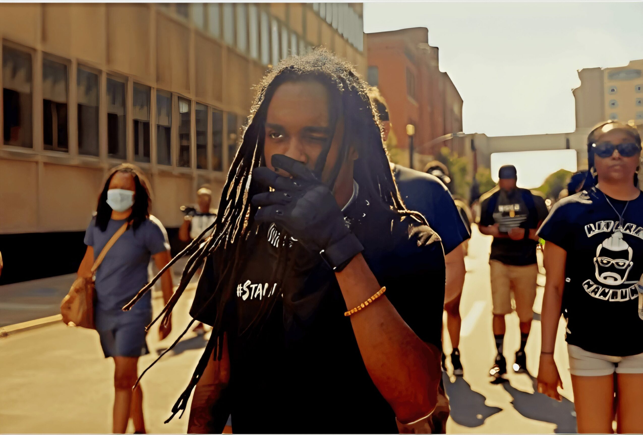 Person with dreadlocks wearing black shirt and gloves, surrounded by people outdoors in sunlight.
