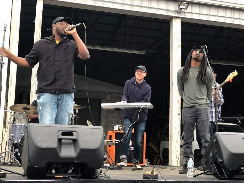Three people perform on an outdoor stage with musical equipment, including a keyboard and microphones, under a roof structure.