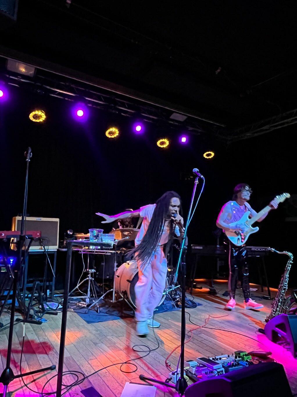 Musicians perform on stage with instruments and equipment, illuminated by purple and yellow stage lights, in a dark venue.