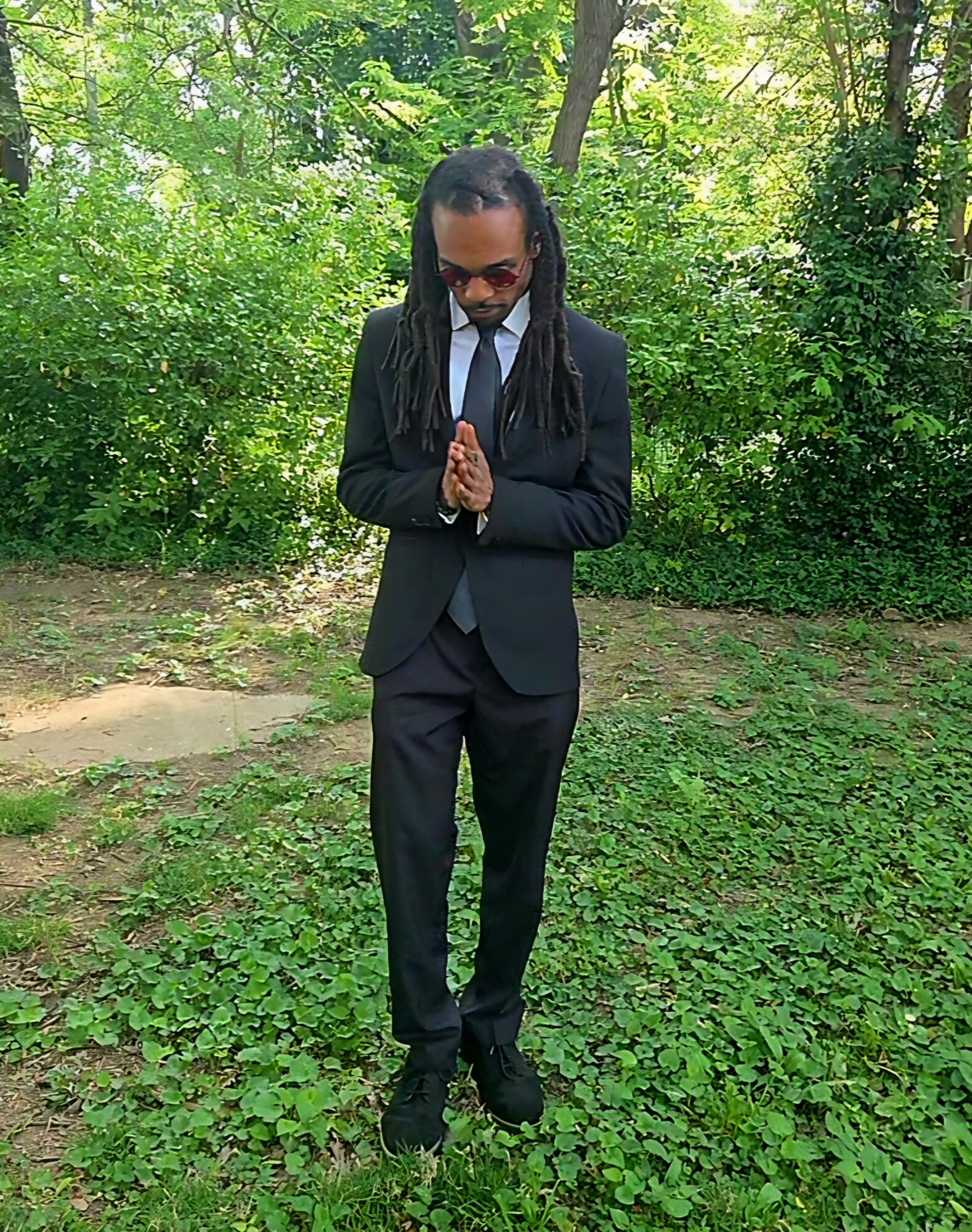 Person in a black suit with hands together, standing outdoors on grass with trees in background.