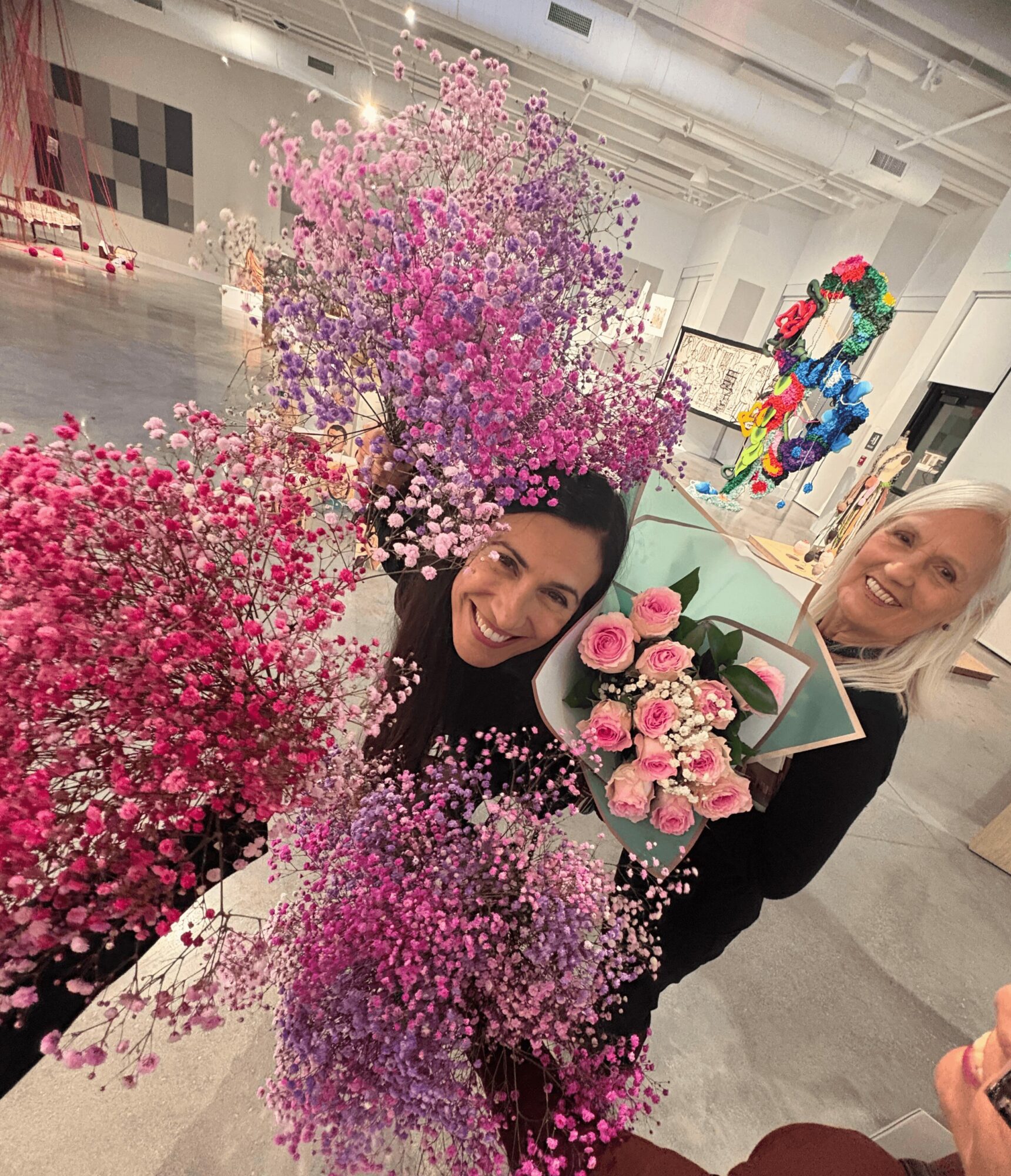 Two women smiling with pink and purple flowers in a spacious indoor setting.
