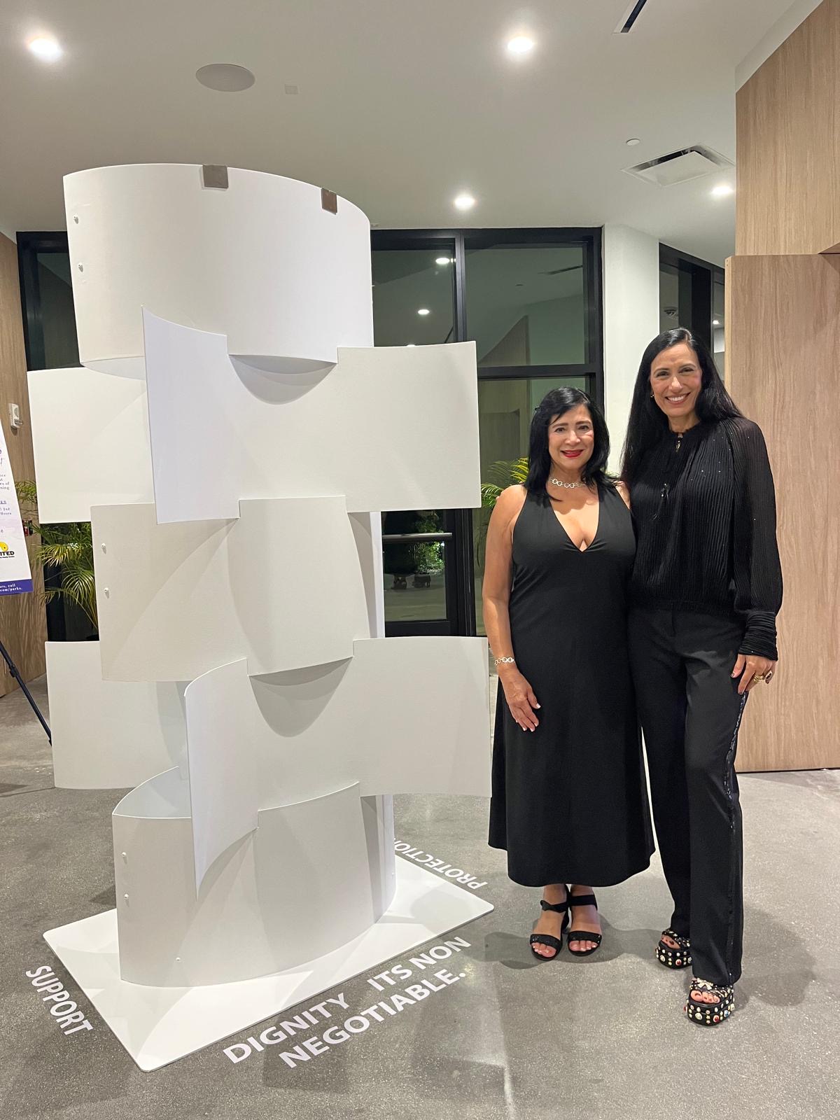 Two women stand side by side indoors, smiling, with a white abstract sculpture behind them and a modern interior setting.