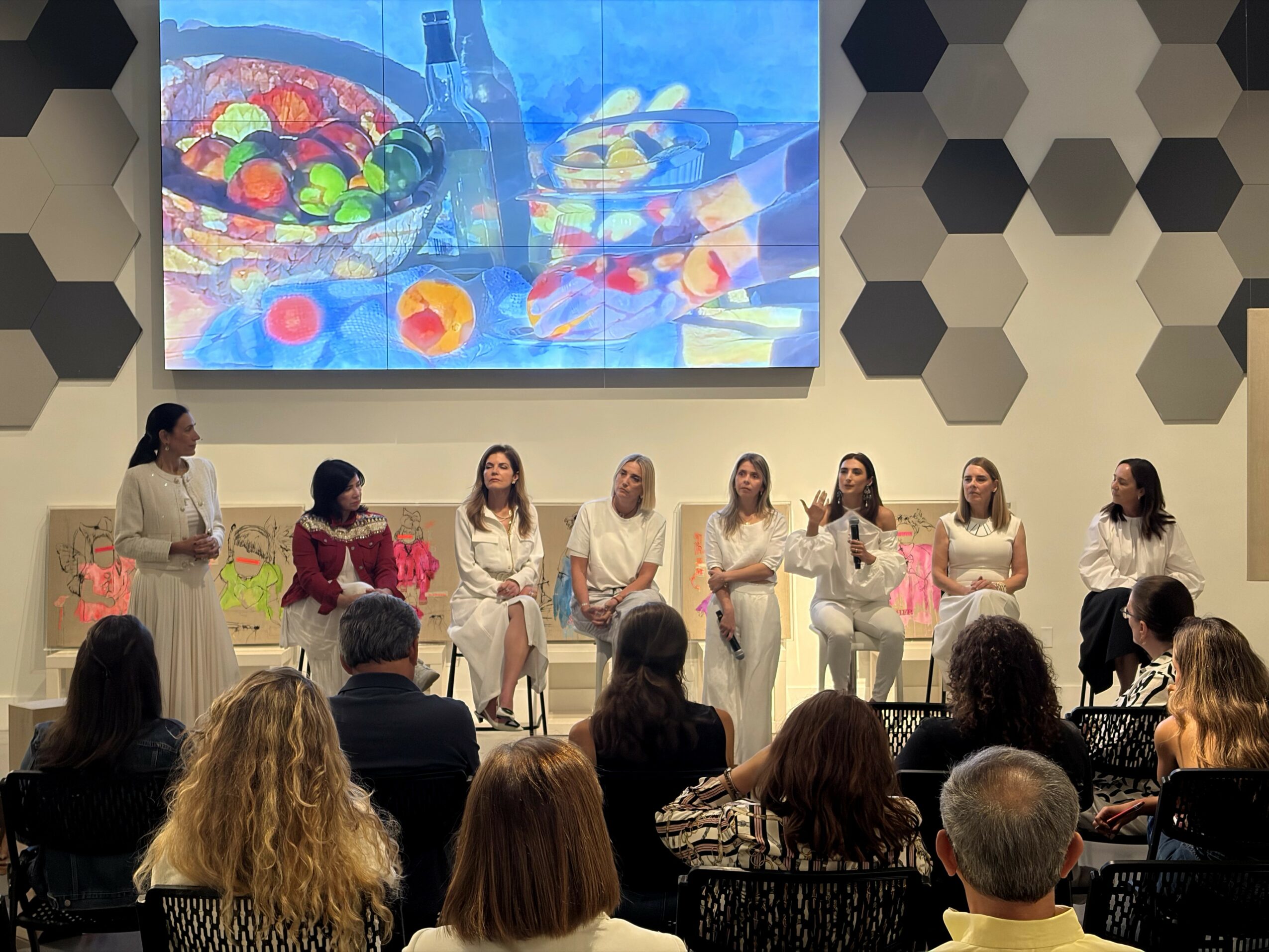 Seven women sit on stage in front of an audience, with a large screen displaying colorful objects behind them.