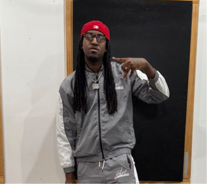Man with long hair, glasses, red cap, gray and white tracksuit, pointing at shoulder, standing indoors.