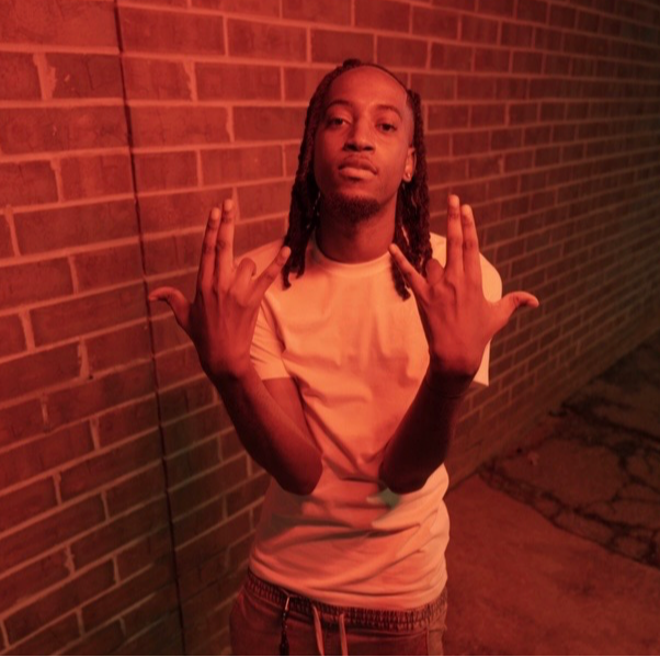 Young person with dreadlocks standing against a brick wall, making hand gestures, wearing a light-colored t-shirt.