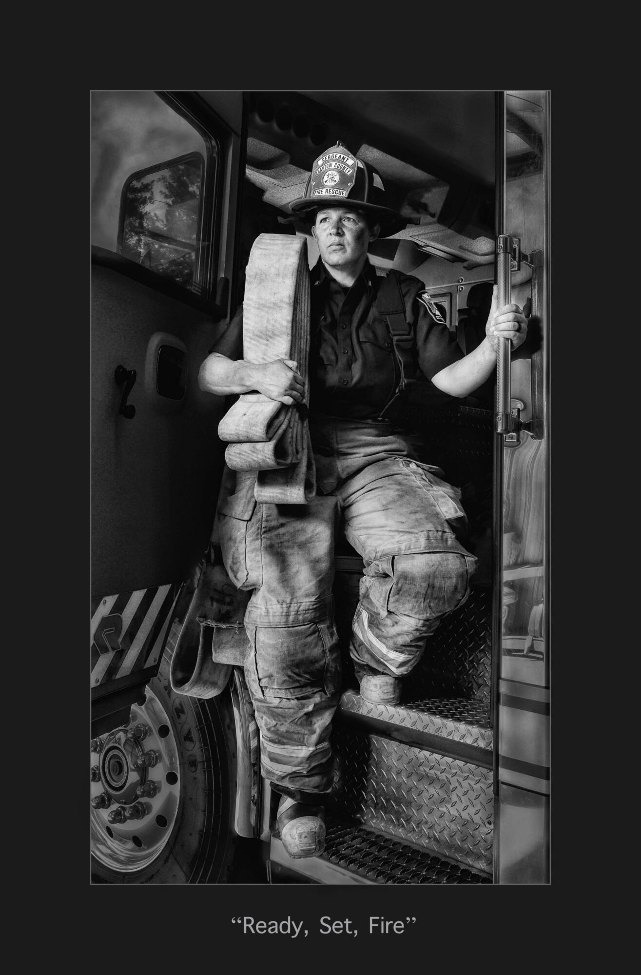 Firefighter in uniform standing at the door of a fire truck, holding a tool, with equipment and a fire scene in the background.