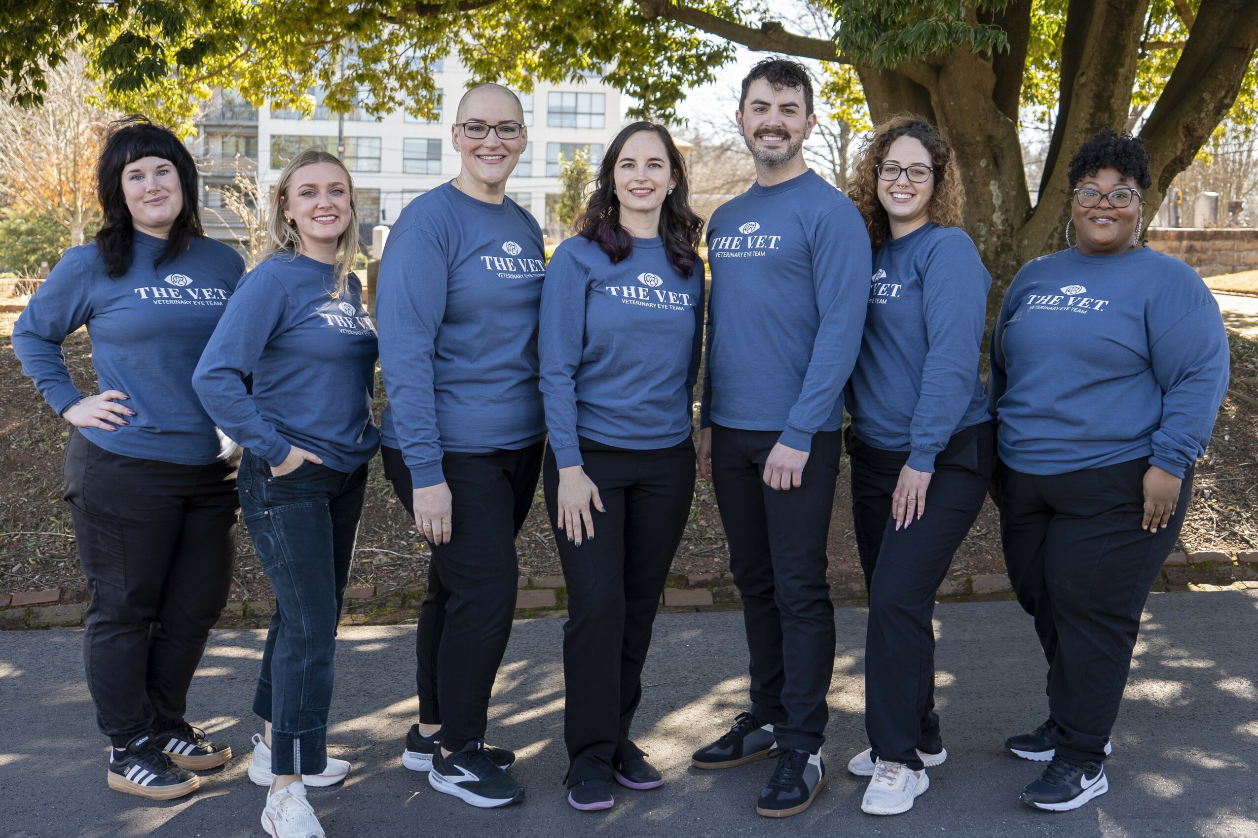 Group of seven people standing outdoors under a tree, wearing matching blue sweatshirts, smiling, with buildings in background.