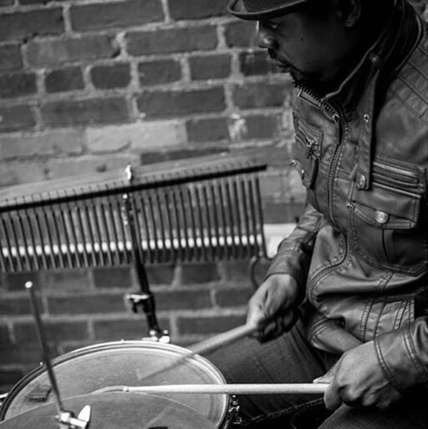 Person playing drums outdoors against a brick wall, wearing a leather jacket and hat, focused on the instrument.