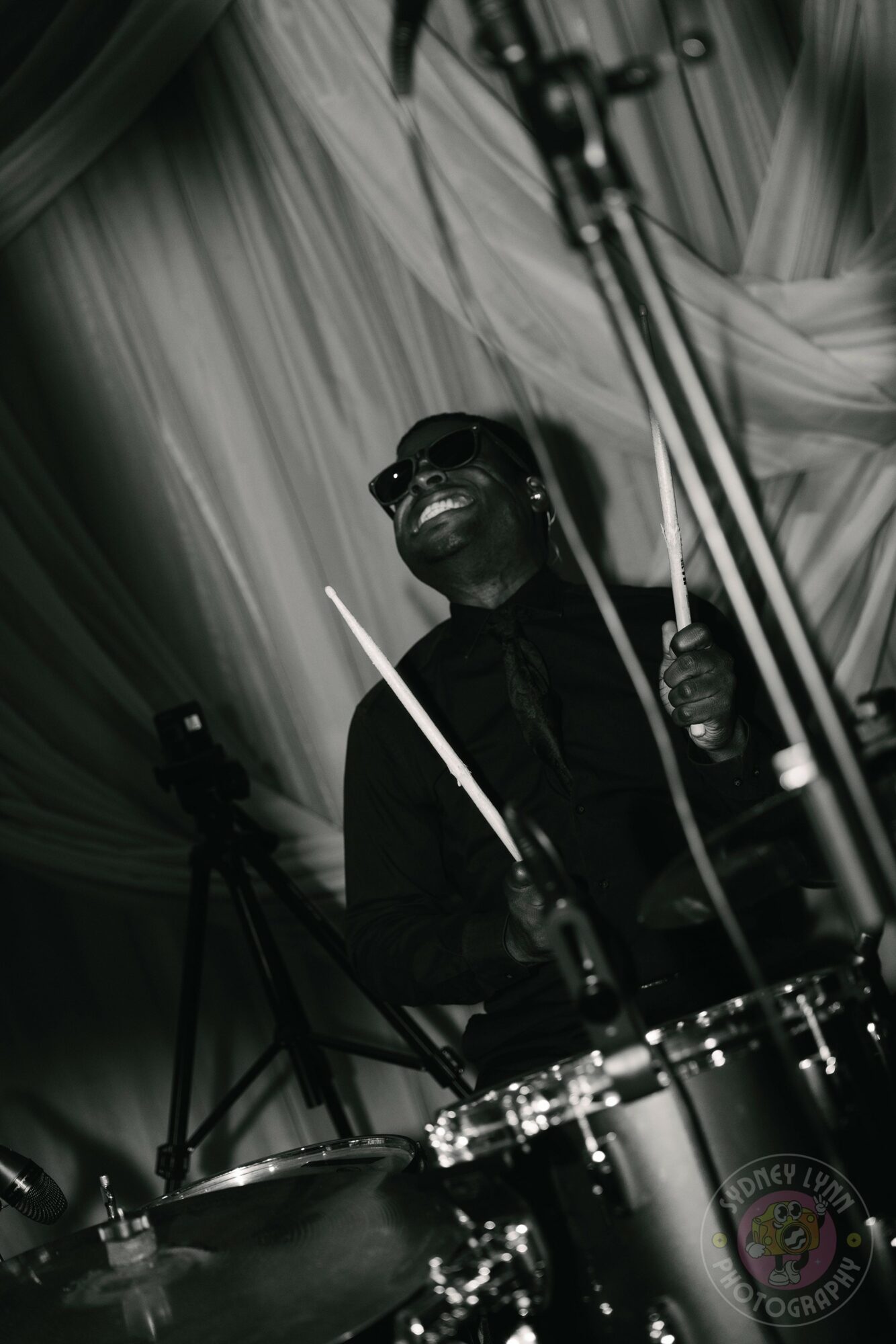 Musician playing drums with drumsticks, smiling, in a stage setting with curtains and equipment.