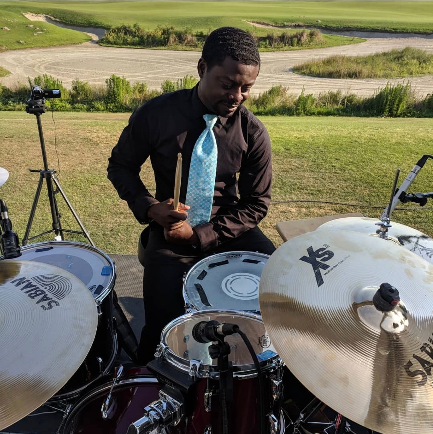 Man in black shirt and blue tie adjusting drum set outdoors with grass and trees in background.