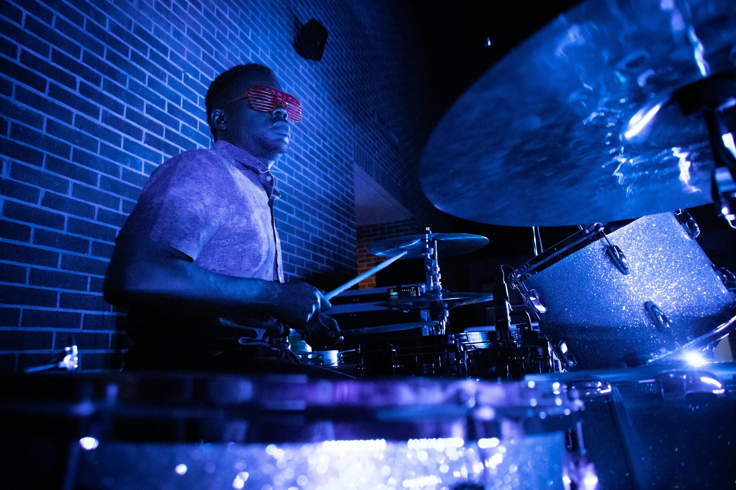 Person playing drums in a dimly lit room with blue lighting and brick wall background.