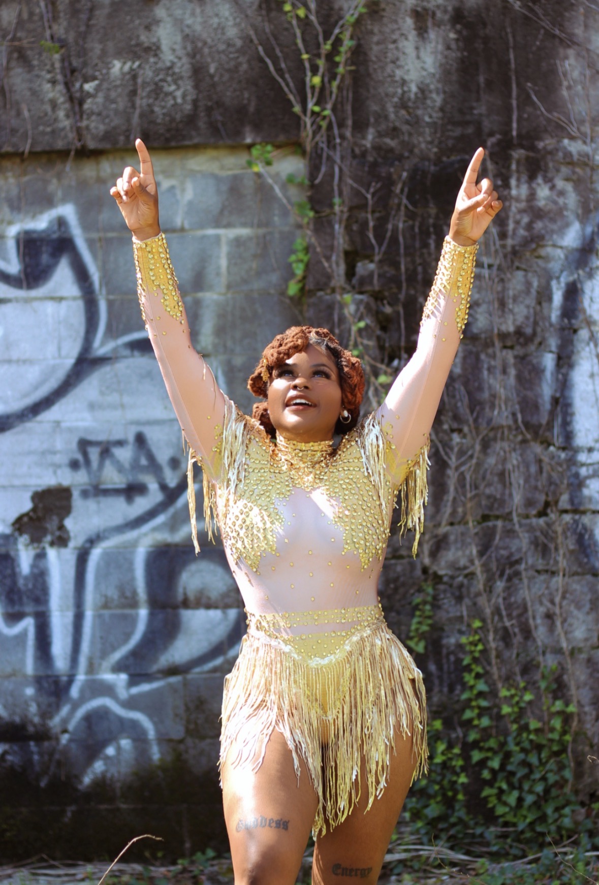 Person in a gold and white costume with arms raised outdoors against a graffiti wall.