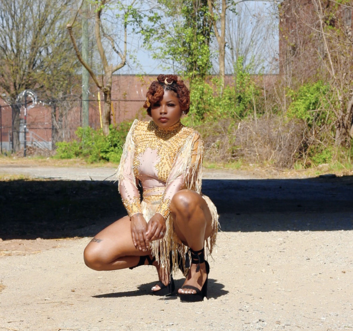 Woman crouching outdoors with trees and a brick wall in background, wearing a gold and pink dress and black heels.