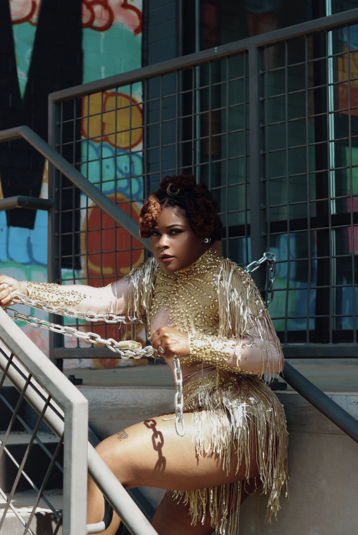 Woman in gold dress sitting on stairs holding a chain, colorful background with grid and painted circles.