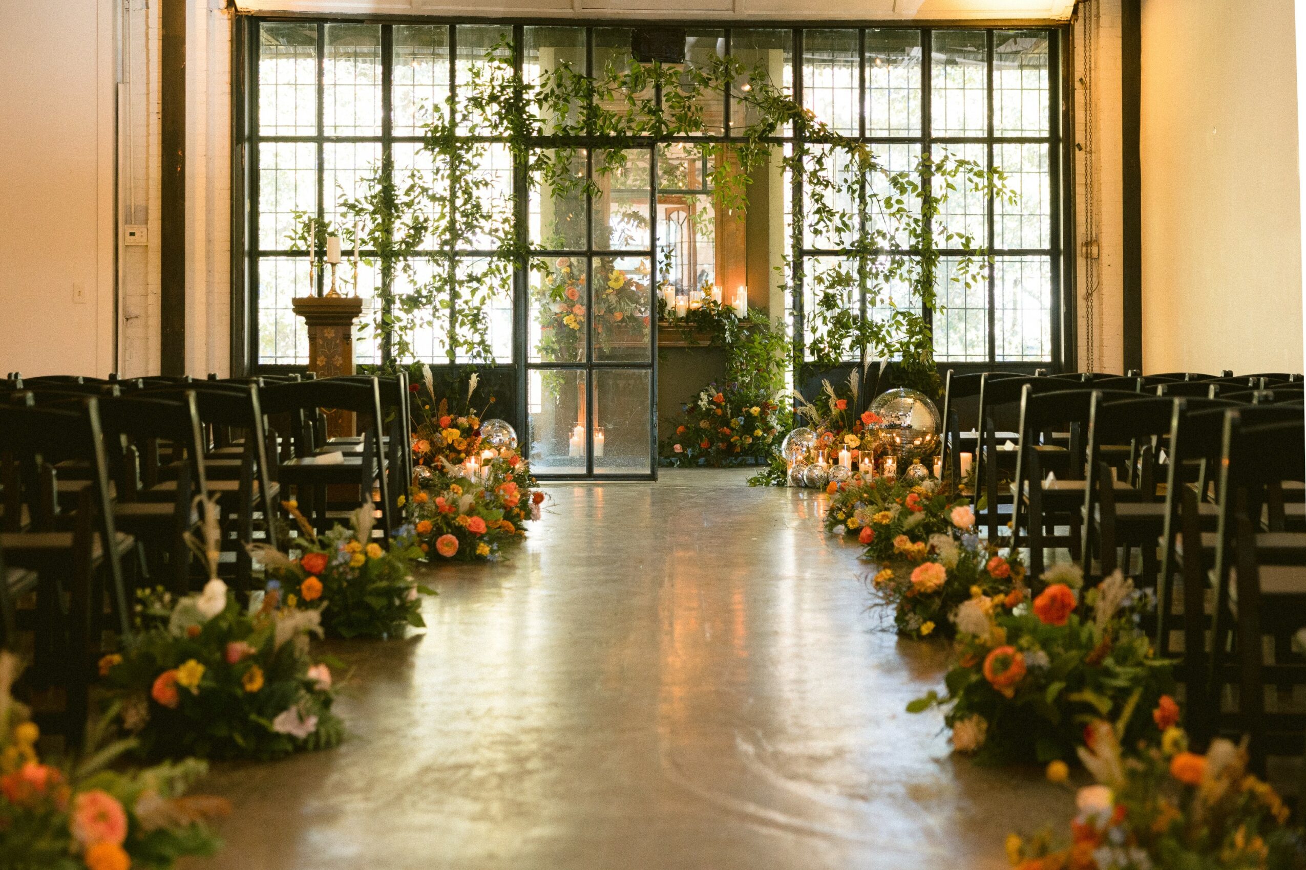 Indoor event space with chairs arranged on both sides, floral arrangements along the aisle, large window with plants outside, and a door at the end.