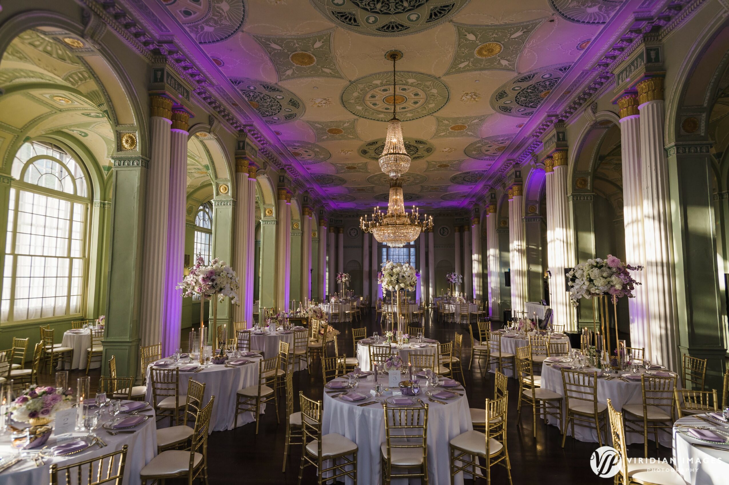 Elegant banquet hall with chandeliers, tall windows, decorated tables, and floral centerpieces, illuminated with purple lighting.