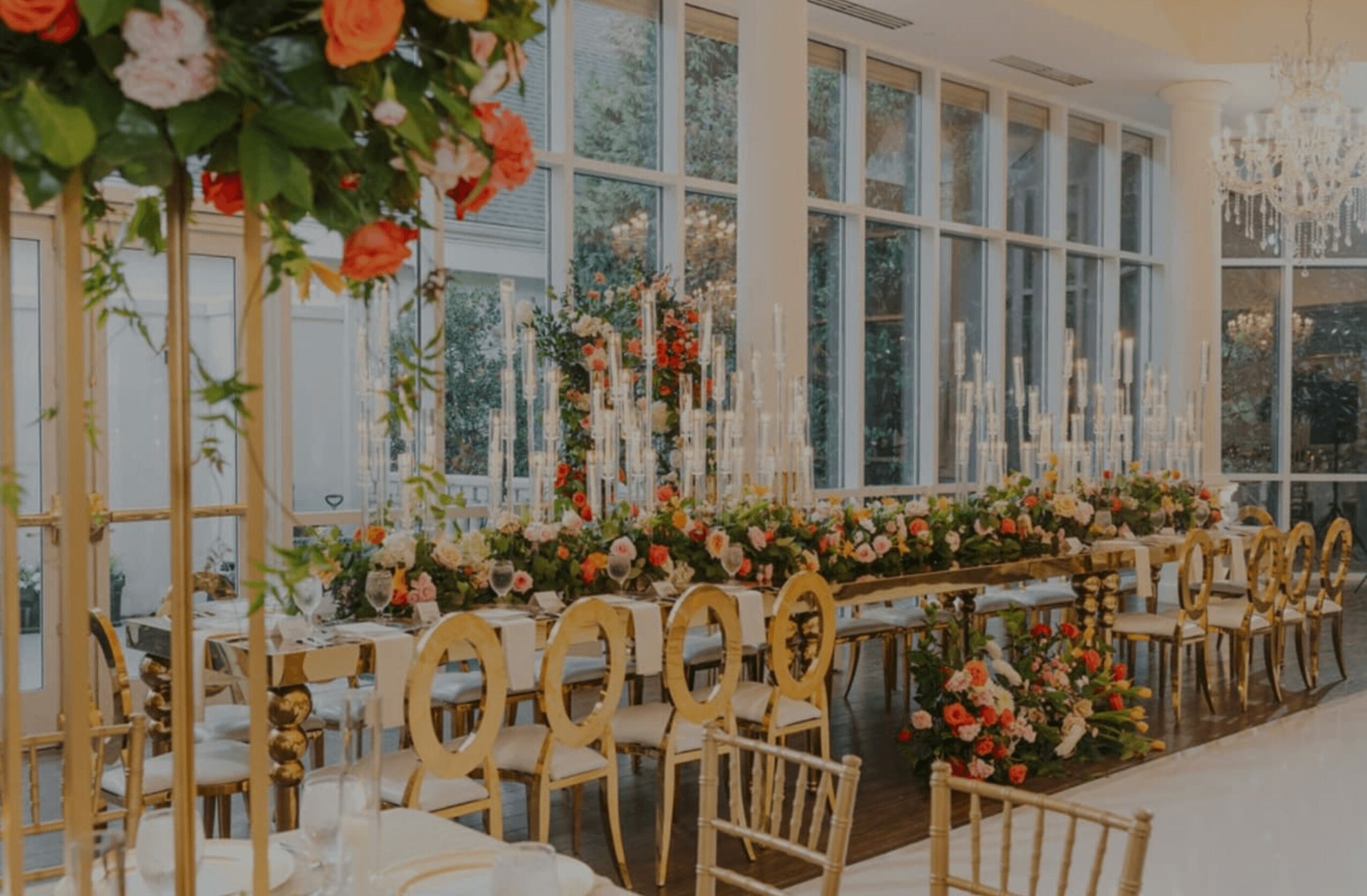 Elegant indoor event space with large windows, floral arrangements, and gold chairs arranged along a table.