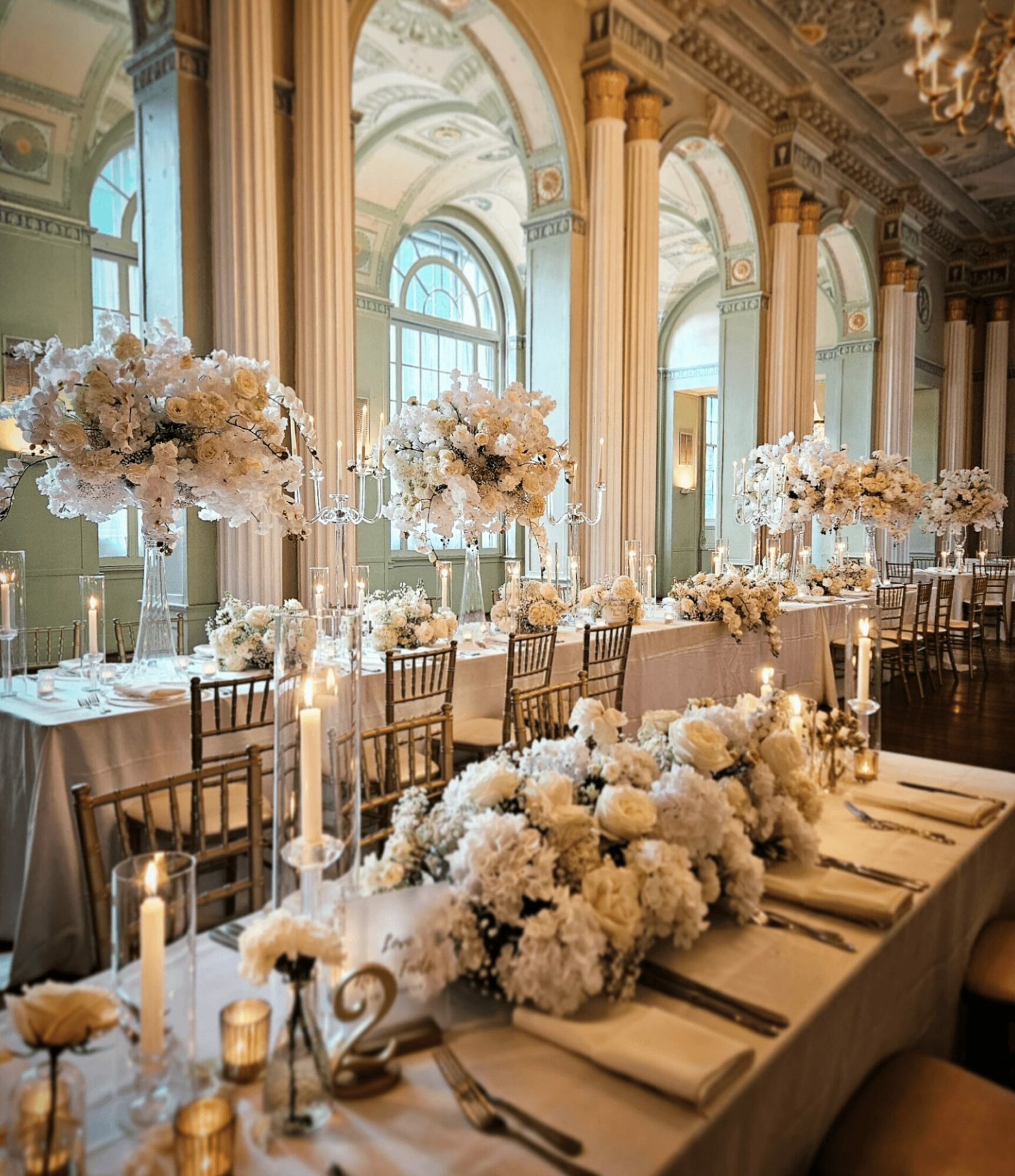 Elegant banquet table with floral arrangements, candles, and place settings in a grand hall with tall windows and columns.