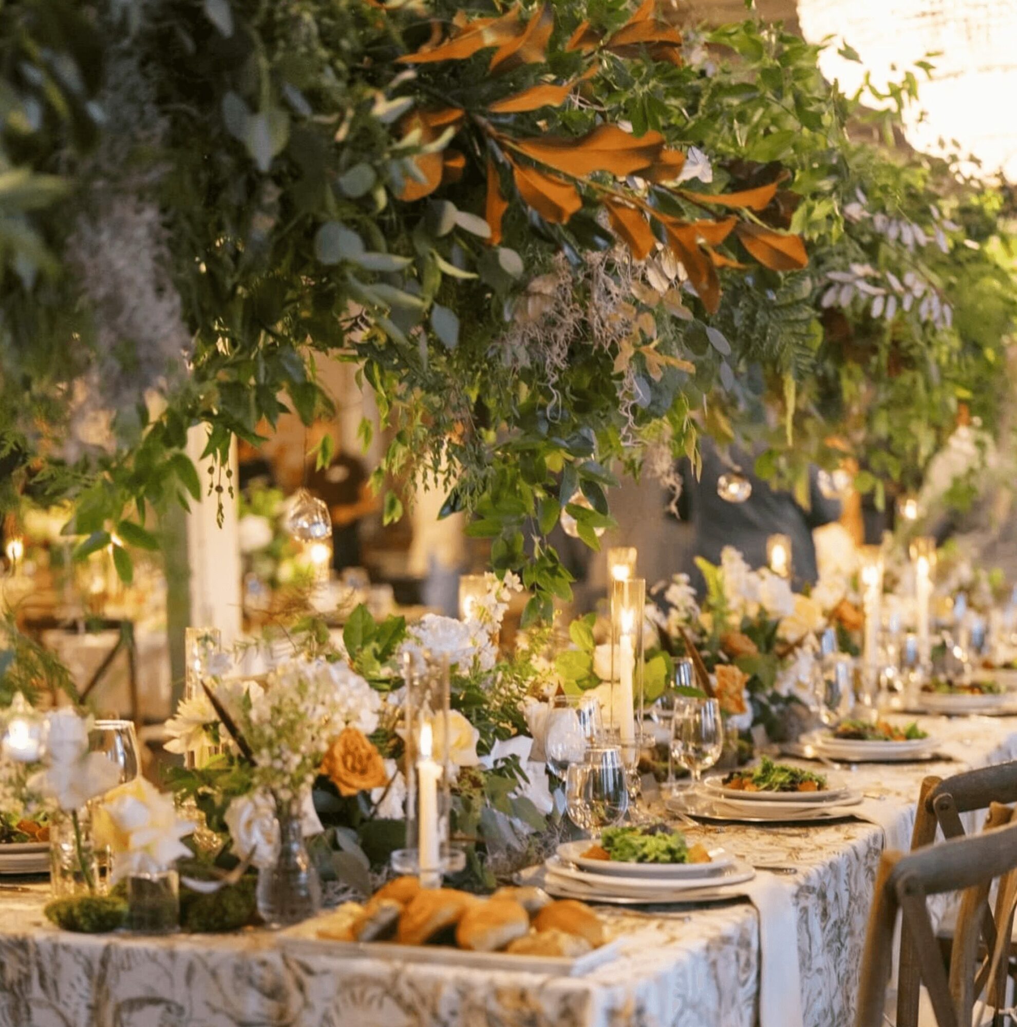 Long decorated table with flowers, candles, and place settings outdoors, surrounded by greenery and warm lighting.