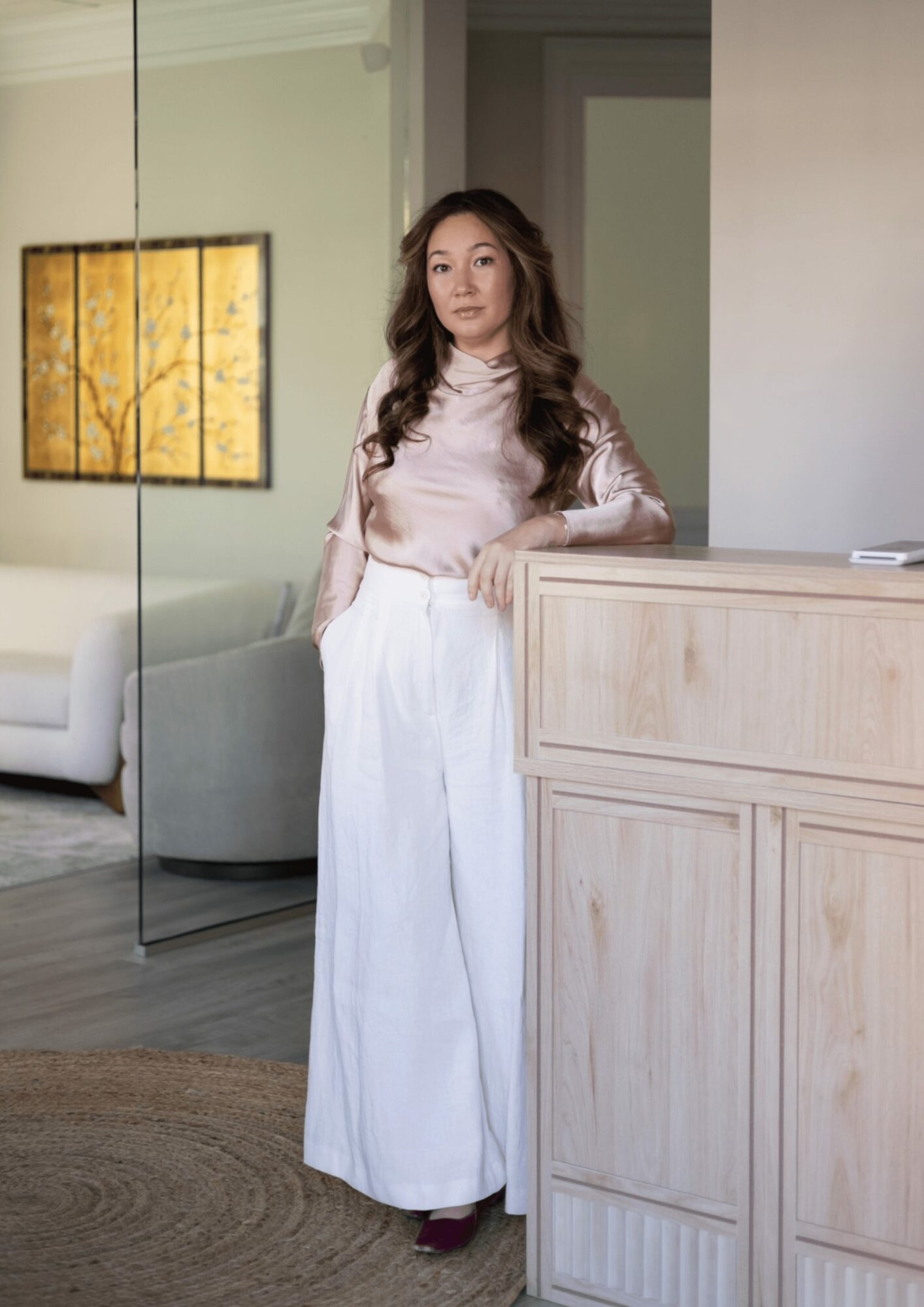 Woman with long wavy hair in pink blouse and white pants standing indoors near a wooden counter.