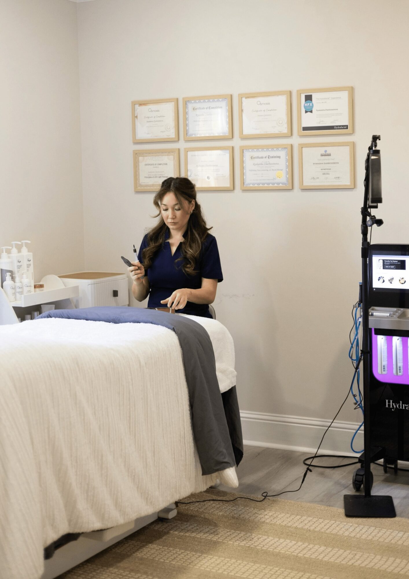 Woman sitting on a bed in a medical or spa setting, looking at her phone, with framed certificates on the wall.